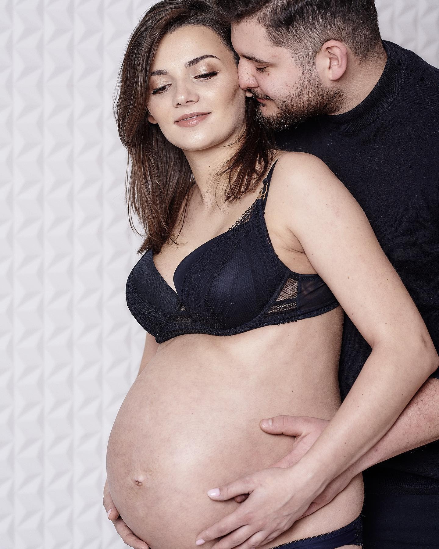 Kobieta w bieliźnie, z dużym ciążowym brzuchem, obejmowana przez mężczyznę w czarnym golfie na tle białej, geometrycznej ściany.