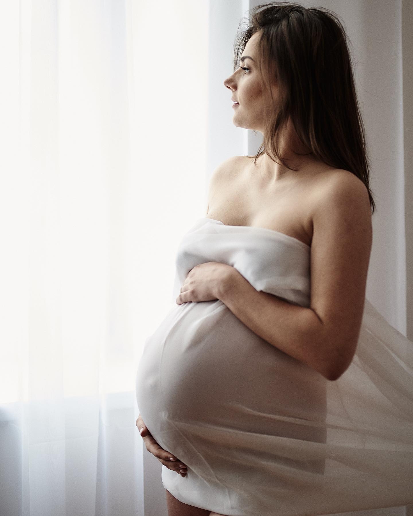 Kobieta w ciąży, owinięta w białą tkaninę, patrzy przez okno w jasnym świetle.