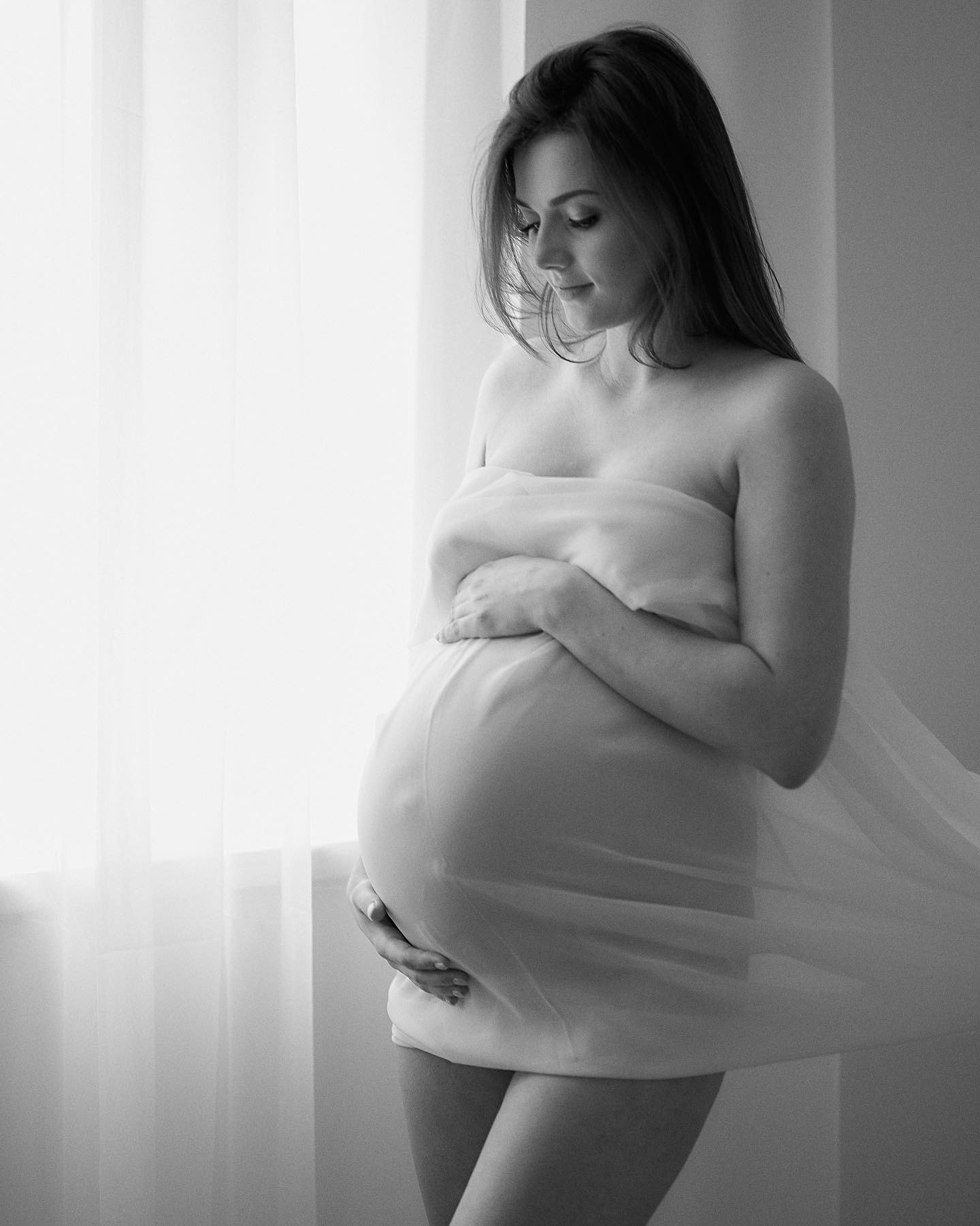 Czarno-białe zdjęcie kobiety w ciąży, owiniętej w jasną tkaninę, stojącej przy oknie z delikatnymi firanami, zamyślony wzrok skierowany w dół.