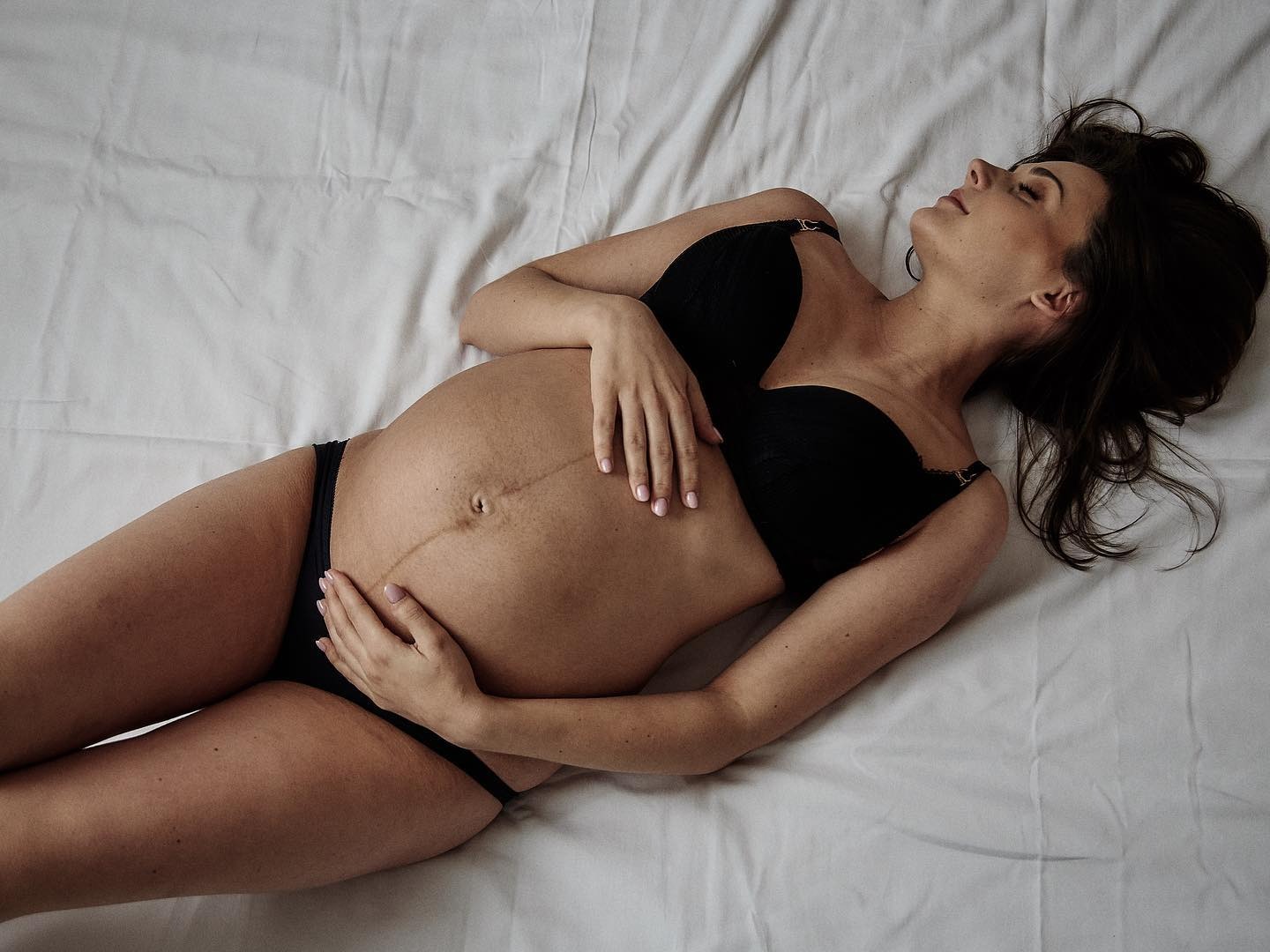 Kobieta w czarnej bieliźnie leży na białym prześcieradle, dłońmi obejmuje ciążowy brzuch, z zamkniętymi oczami i rozpuszczonymi włosami.