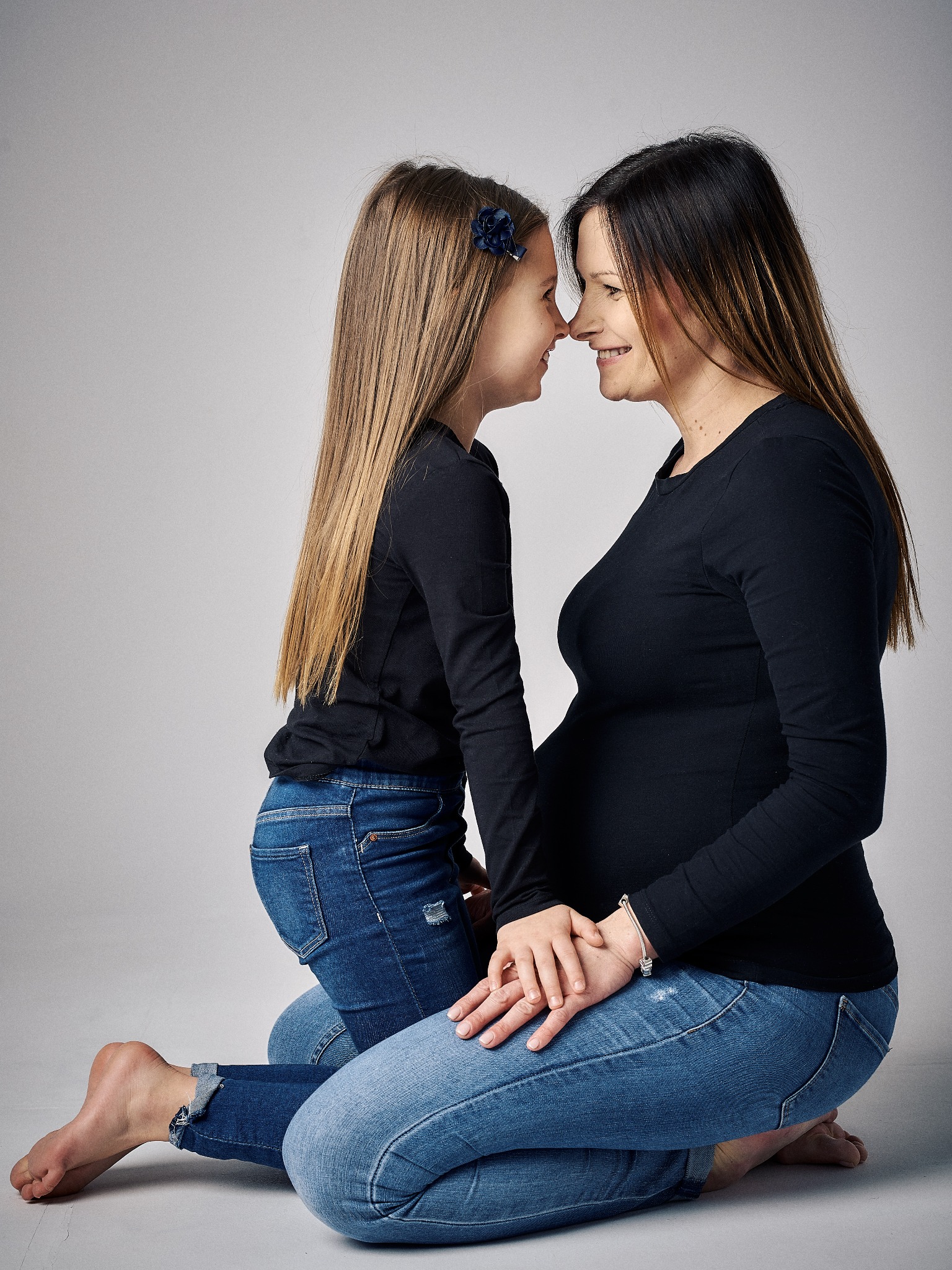 Czuła sesja ciążowa: Uśmiechnięta, ciężarna mama i jej córka dotykają się nosami, siedząc na kolanach na jasnym tle, obie ubrane w czarne bluzki i dżinsy.