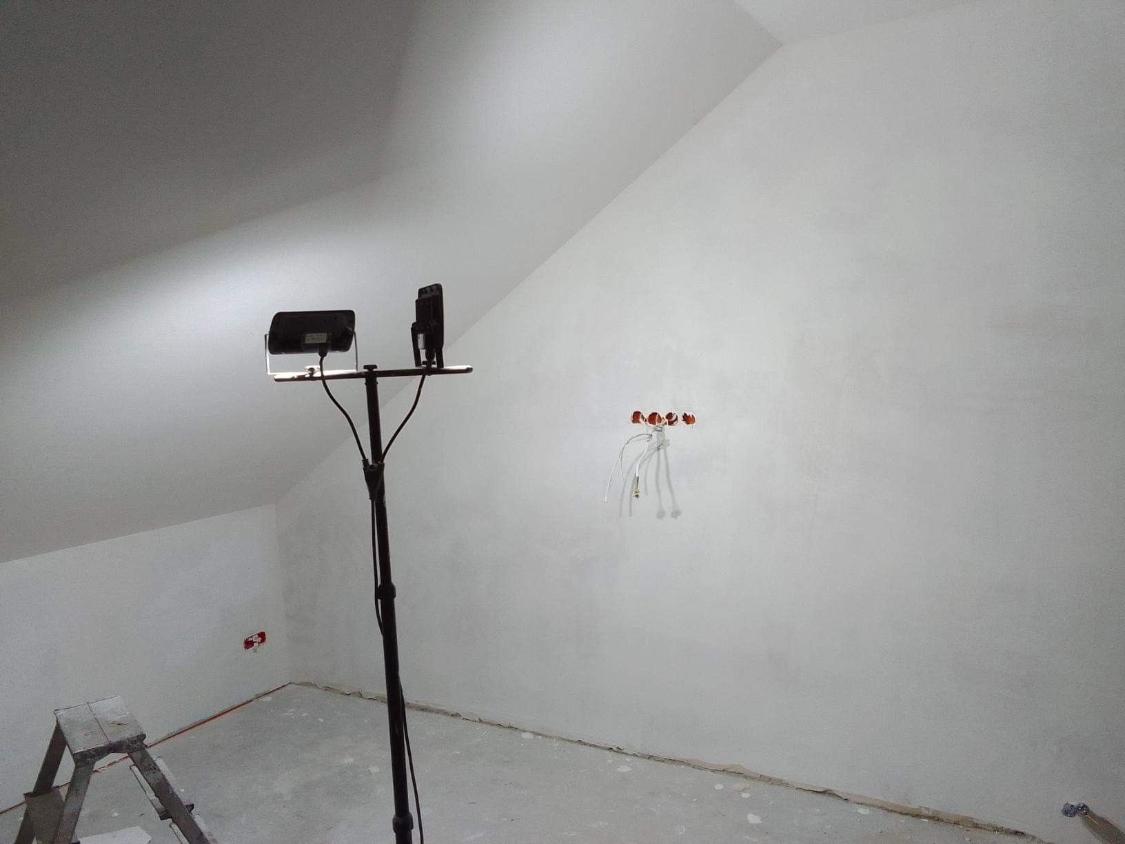 Poddasze w trakcie prac wykończeniowych: świeżo zagruntowane ściany, widoczne puszki elektryczne z wystającymi kablami, oświetlenie budowlane na statywie i drabina malarska w rogu.