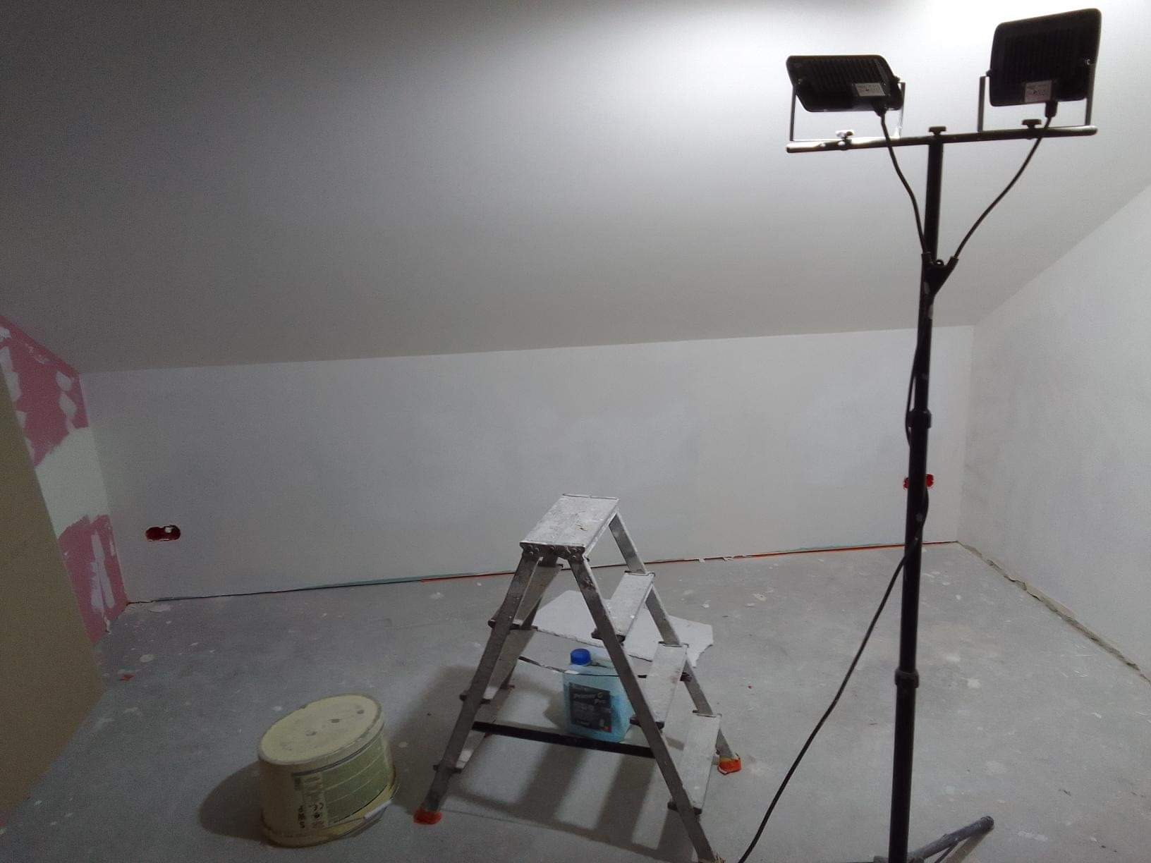 Pokój w trakcie remontu: ściany po szpachlowaniu, drabina z preparatem gruntującym, lampa budowlana oświetlająca sufit pod skosem, ślady po łatach na ścianie.