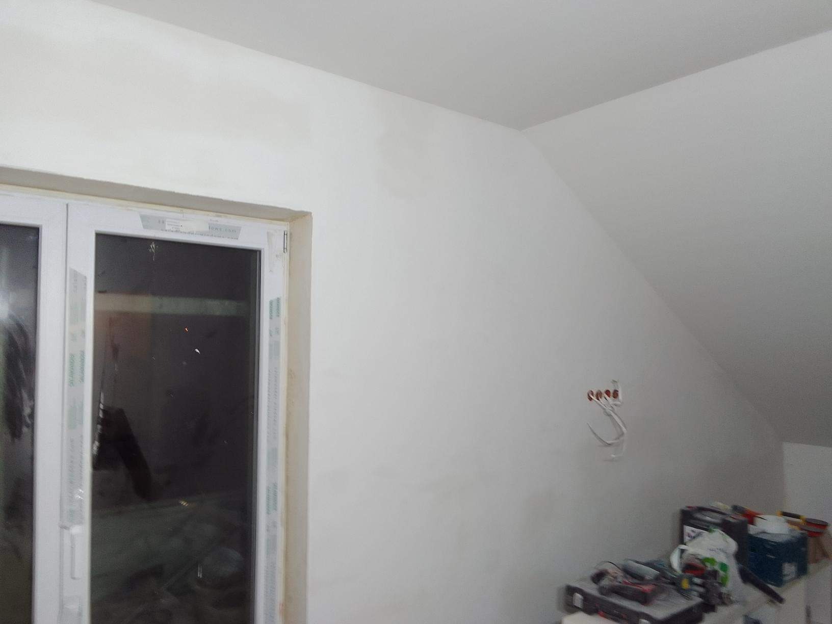 Poddasze po szpachlowaniu, widoczne gładkie, białe ściany, okno PCV oraz wystające kable elektryczne i narzędzia na podłodze.