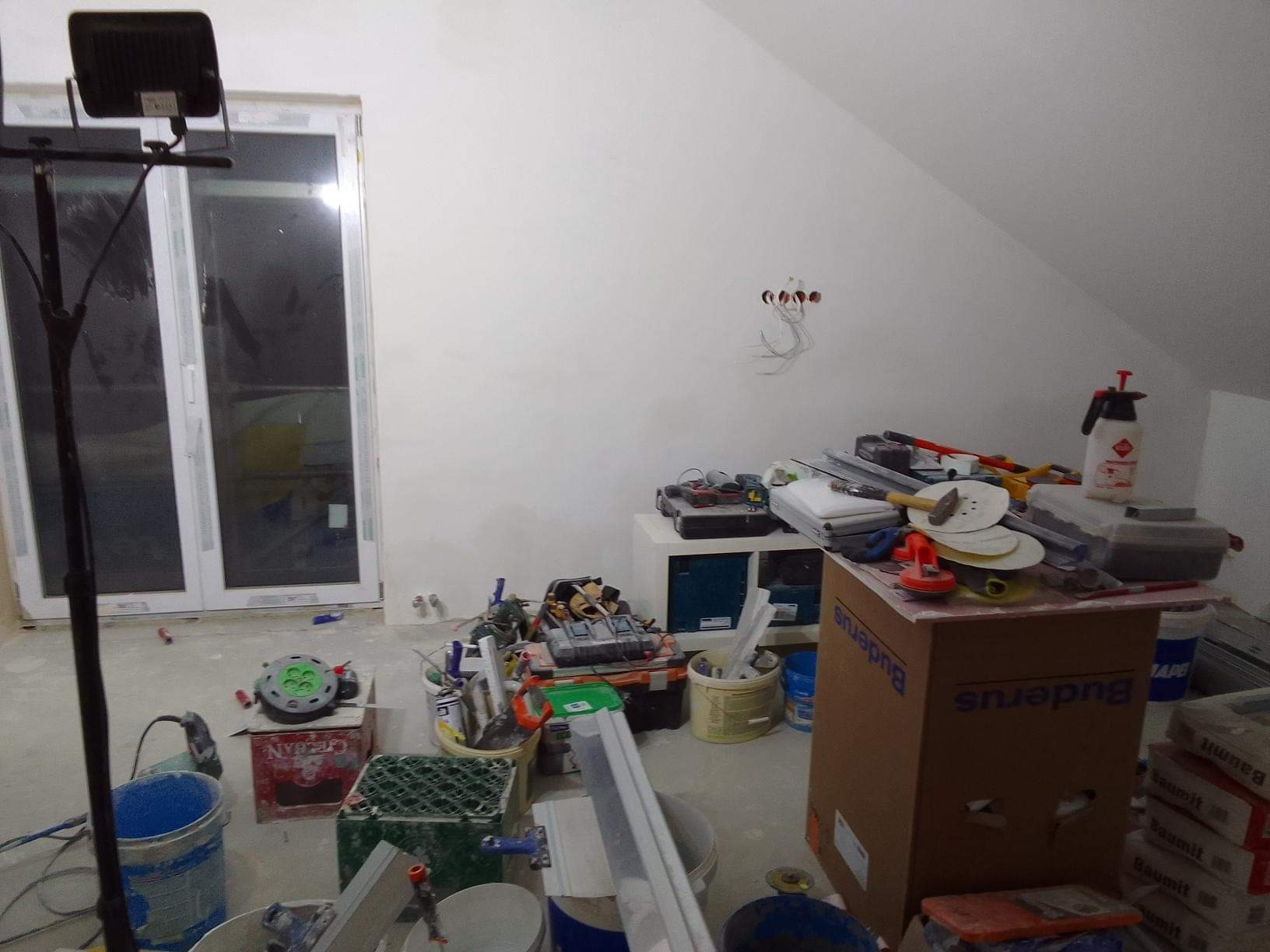 Poddasze w trakcie prac wykończeniowych: gładkie, białe ściany, okno balkonowe, rozstawiony reflektor, narzędzia i materiały budowlane w nieładzie, widoczne puszki instalacyjne bez osprzętu.