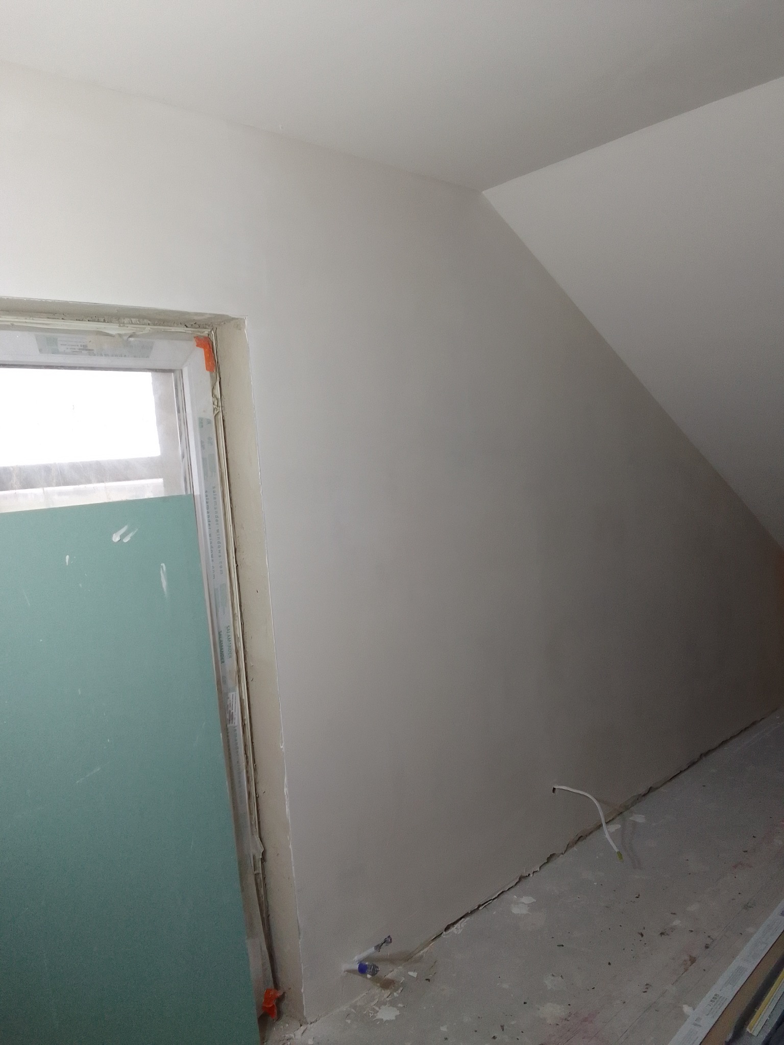 Poddasze po szpachlowaniu, widoczne ściany skośne, okno z zabezpieczeniem folią oraz wystające przewody elektryczne przy podłodze.