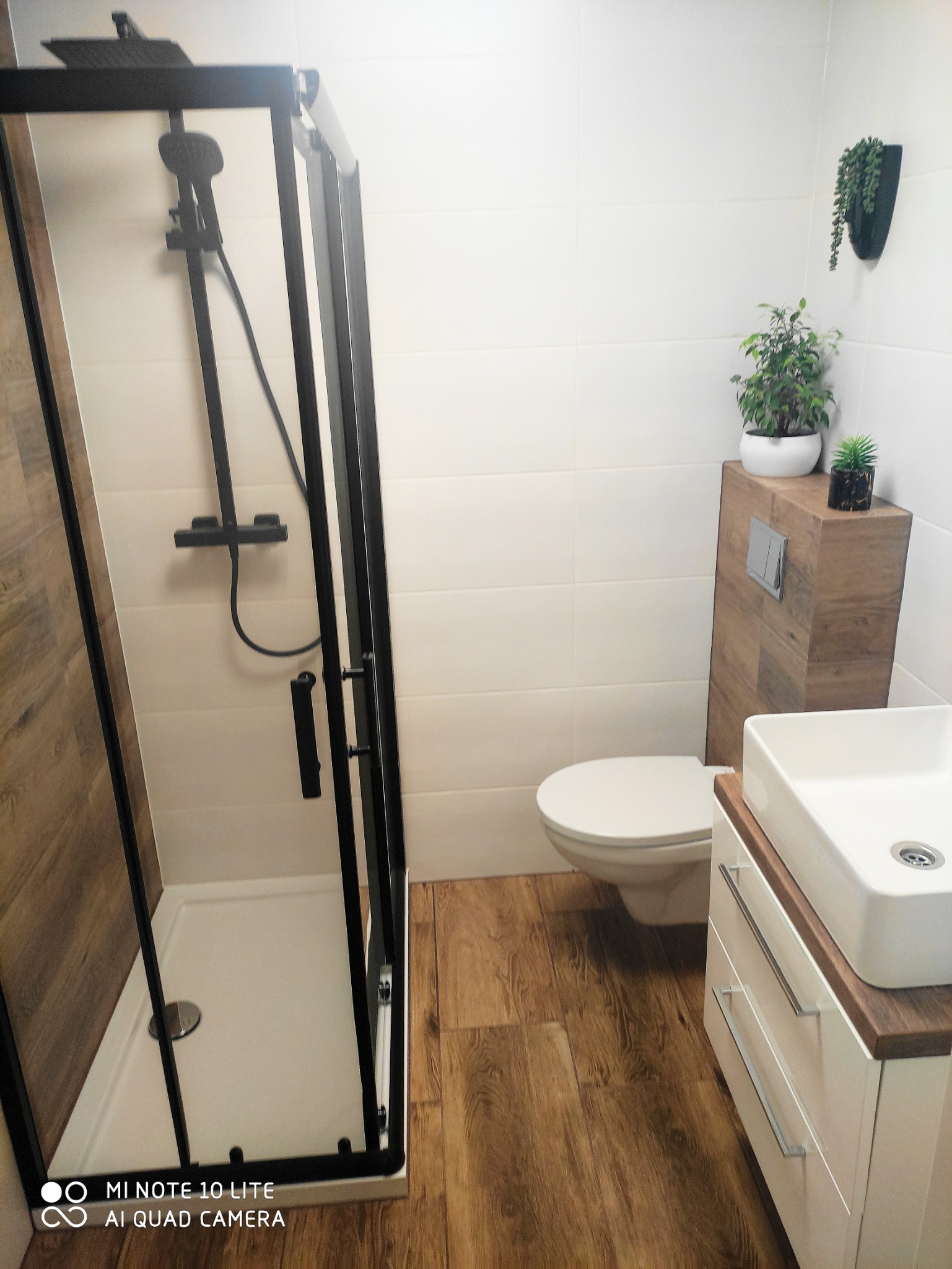 Nowoczesna łazienka z kabiną prysznicową typu walk-in z czarną ramą, umywalką nablatową na szafce z szufladami, toaletą, drewnopodobną podłogą i płytkami imitującymi drewno na obudowie stelaża WC...