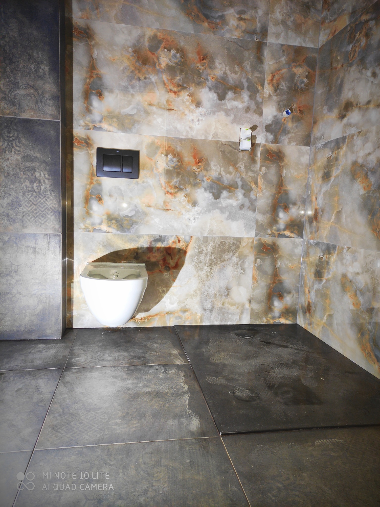 Nowoczesna łazienka z marmurowymi płytkami na ścianach i ciemnymi płytkami podłogowymi, wisząca toaleta z czarnym przyciskiem spłukującym, widoczne ślady po pracach na podłodze.