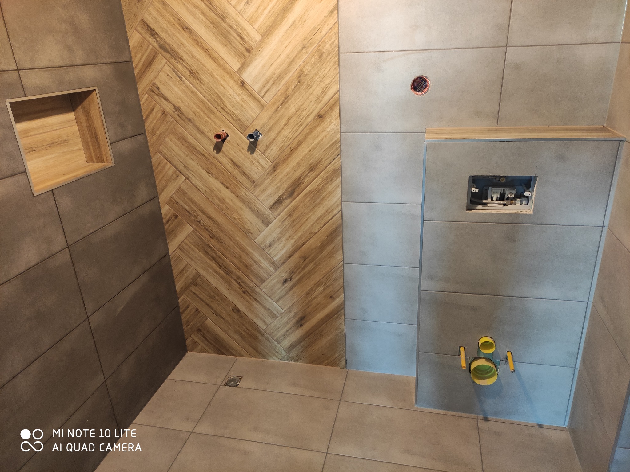 Ściana łazienki wyłożona płytkami w dwóch odcieniach szarości i wzorem drewna ułożonym w jodełkę; widoczne instalacje wodne i kanalizacyjne przed zabudową.