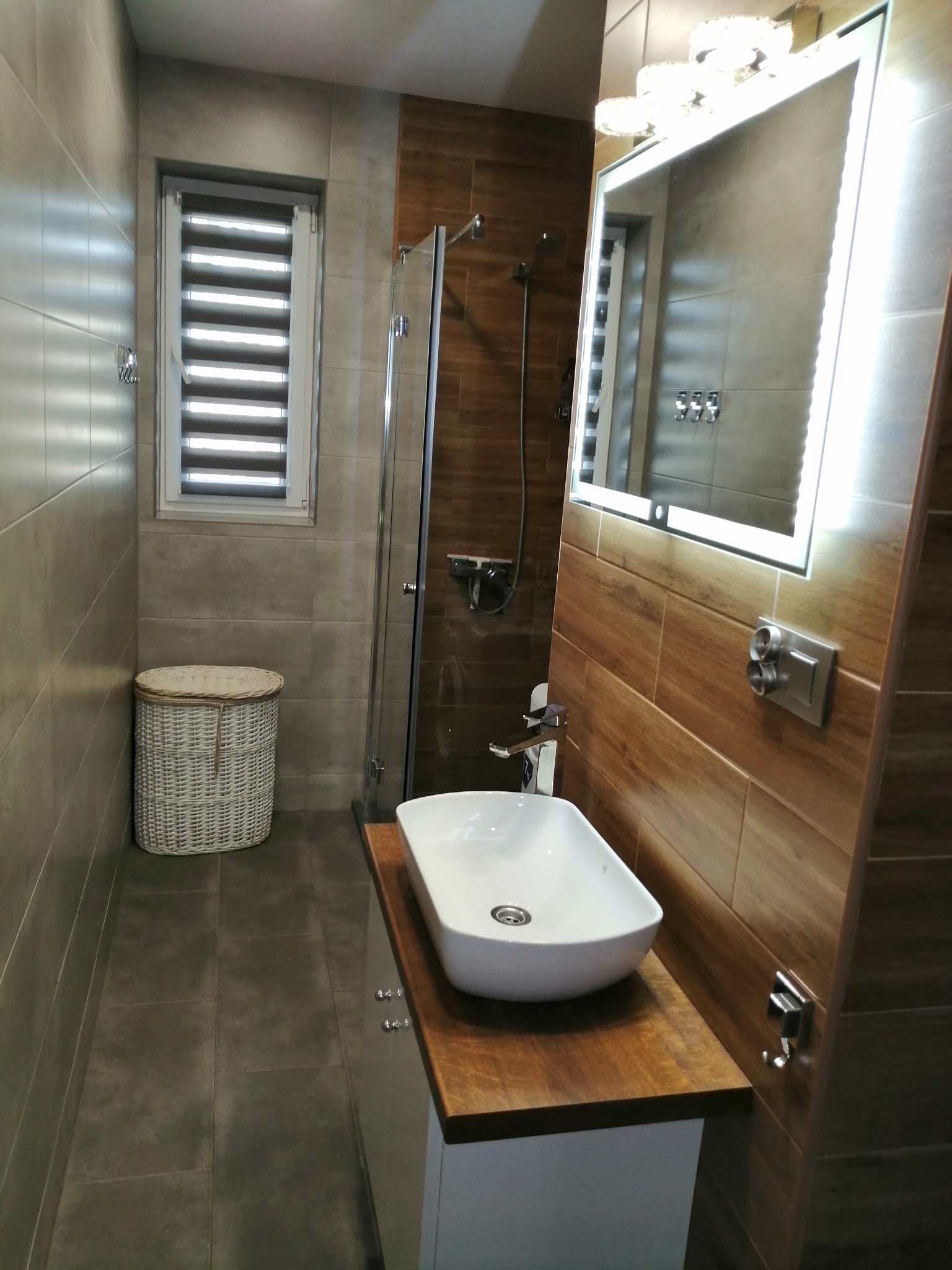 Wykończona łazienka z brązowymi i szarymi płytkami, umywalką nablatową na drewnianym blacie, podświetlanym lustrem i kabiną prysznicową.