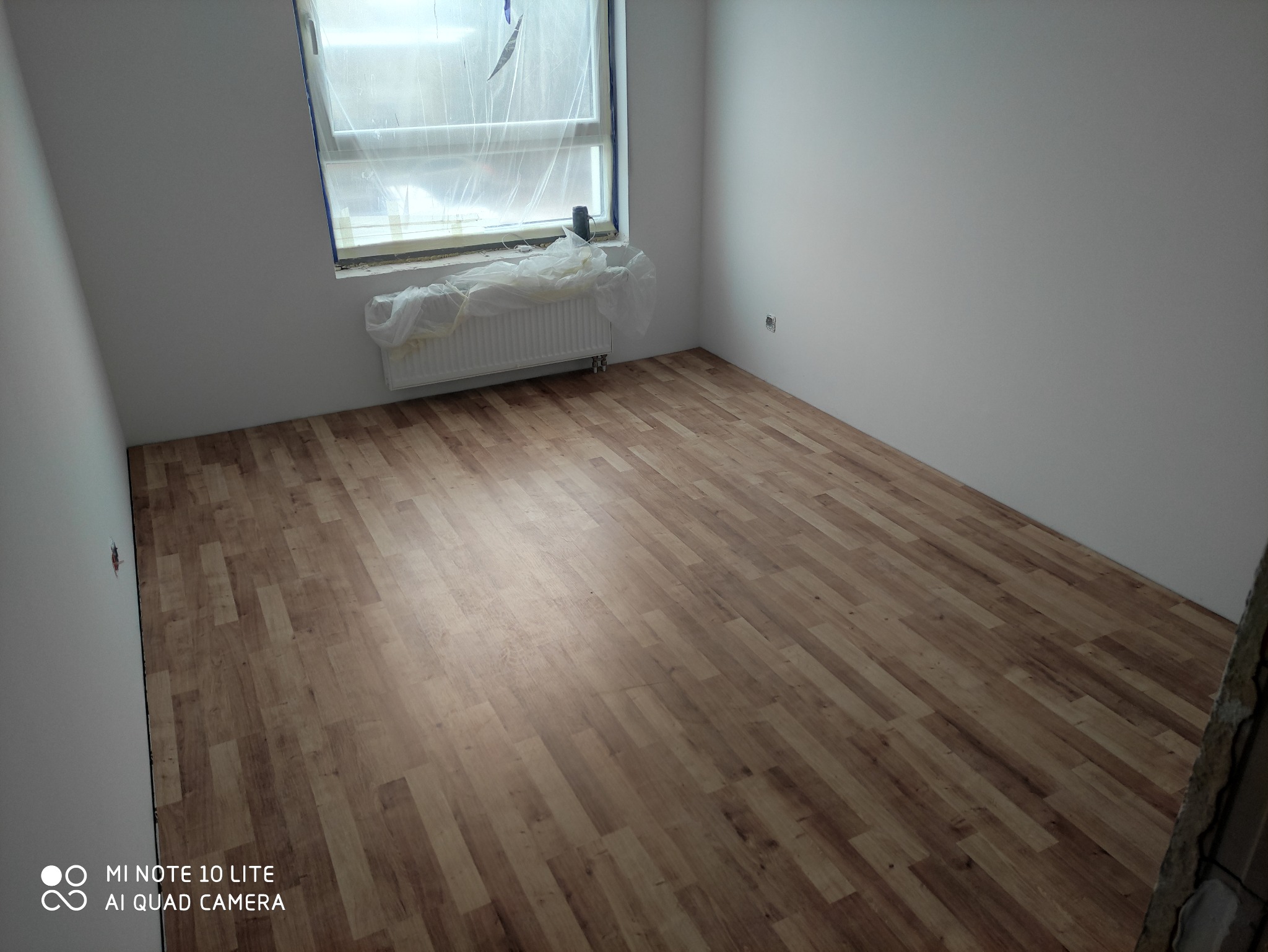 Pokój z nowo ułożonymi panelami podłogowymi w odcieniach brązu, ściany pomalowane na biało, okno zabezpieczone folią malarską, widoczne gniazdko elektryczne i fragment niezakończonego wykończenia...