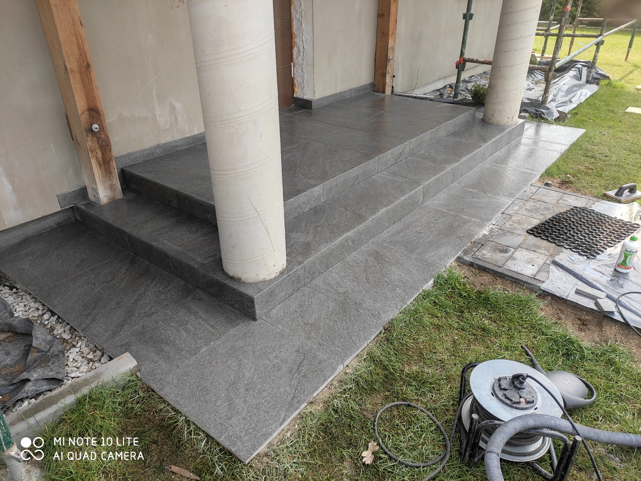 Ułożone szare płytki na schodach wejściowych z kolumnami, widoczne narzędzia i materiały budowlane na trawie obok.