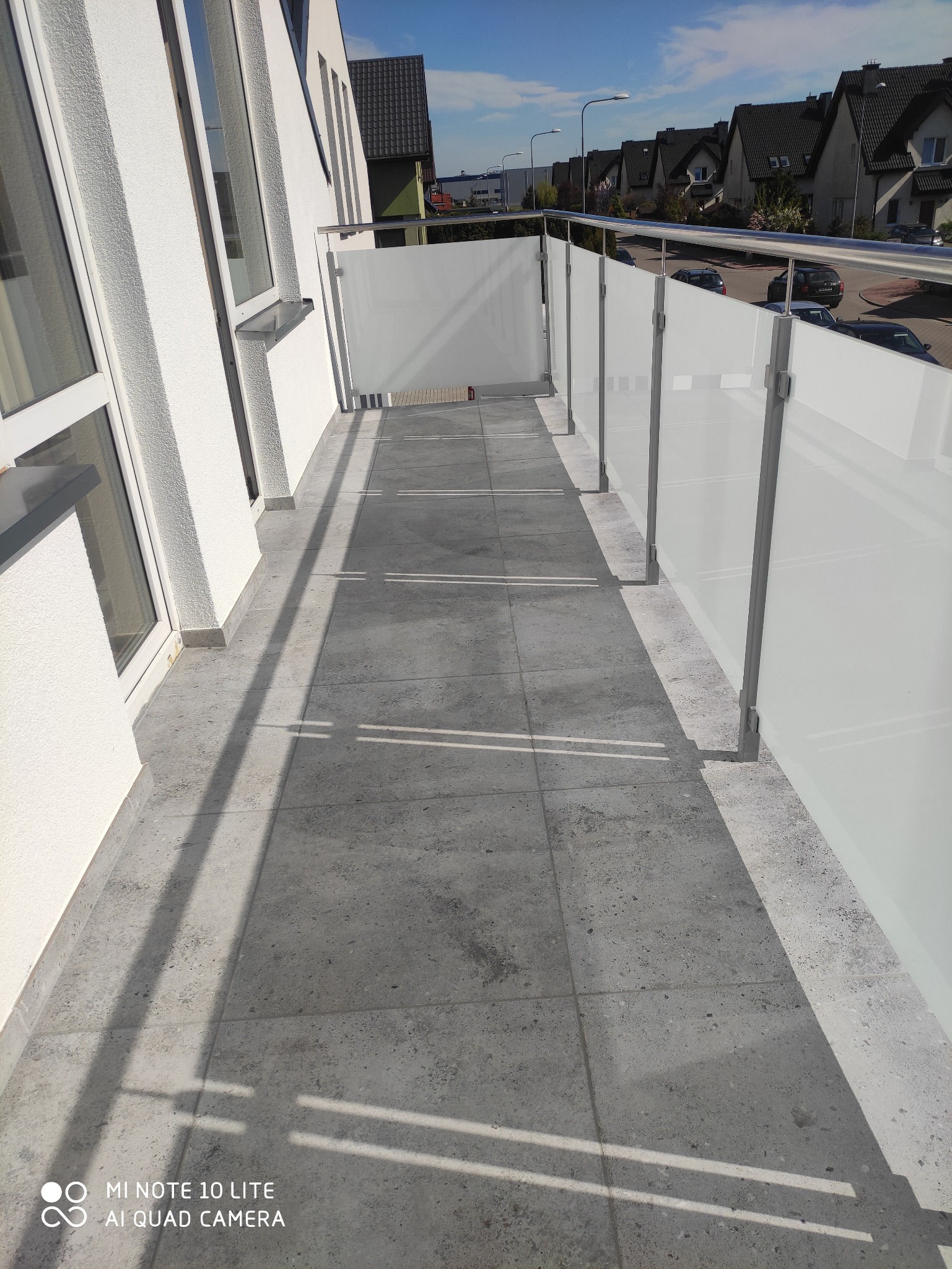 Wyłożony szarymi płytkami balkon z szklaną balustradą i metalowymi poręczami, widok na osiedle domów jednorodzinnych w słoneczny dzień.