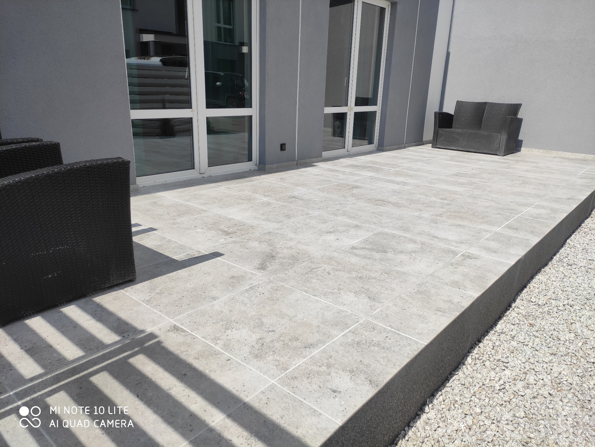 Szary taras wyłożony płytkami z jasnymi fugami, z ciemnymi meblami ogrodowymi i białym żwirem przy krawędzi, widoczne białe ramy okienne i drzwi balkonowych.
