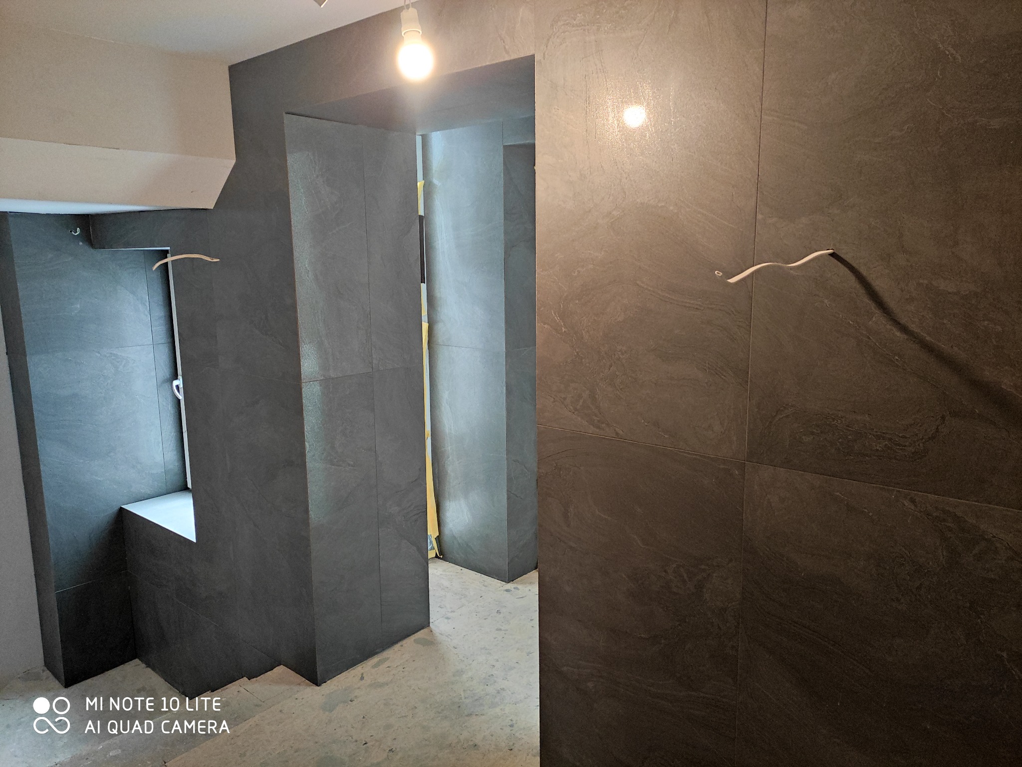 Szare płytki imitujące kamień na ścianach klatki schodowej i w przejściu między pomieszczeniami, widoczny wystający kabel elektryczny i surowy beton na podłodze.