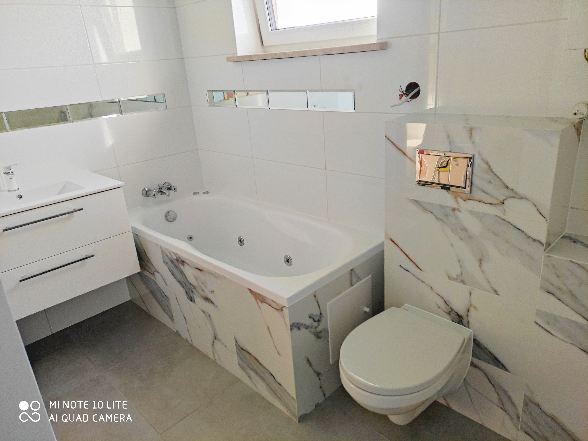 Wykończona łazienka w bieli z akcentami marmuru: wanna z hydromasażem, podwieszana toaleta z chromowanym przyciskiem, umywalka z szafką i lustrzane detale na ścianie.