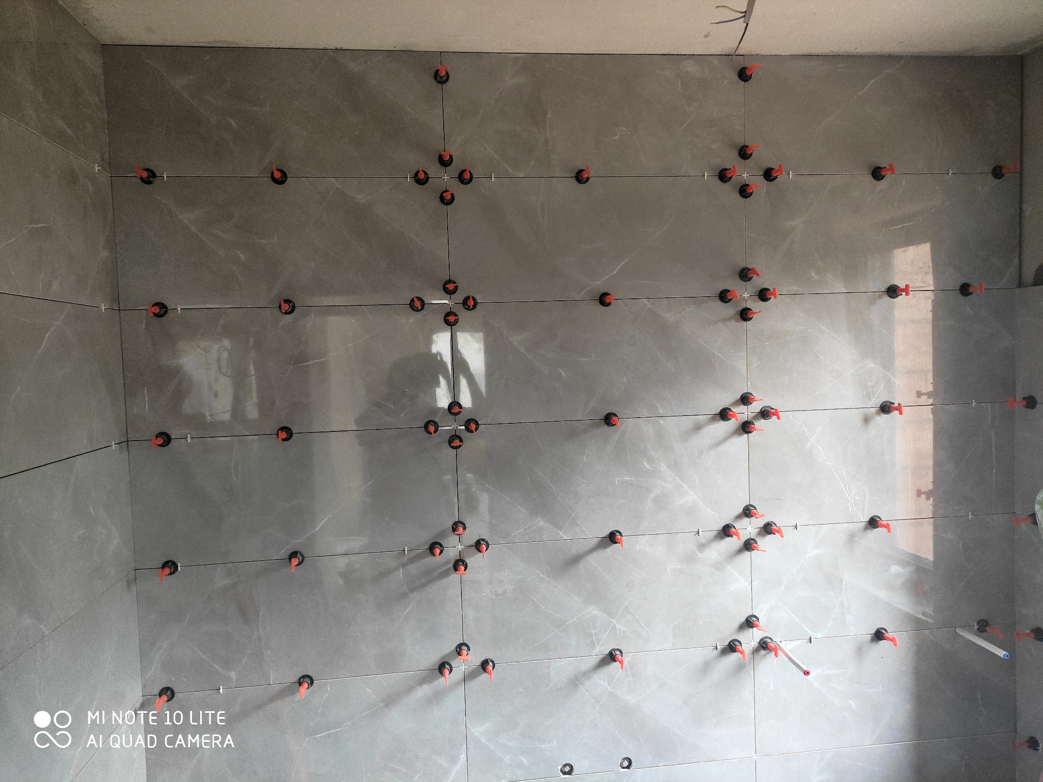 Ściana w trakcie układania płytek ceramicznych w odcieniach szarości z użyciem systemu poziomowania płytek z pomarańczowymi klinami i czarnymi podstawkami.