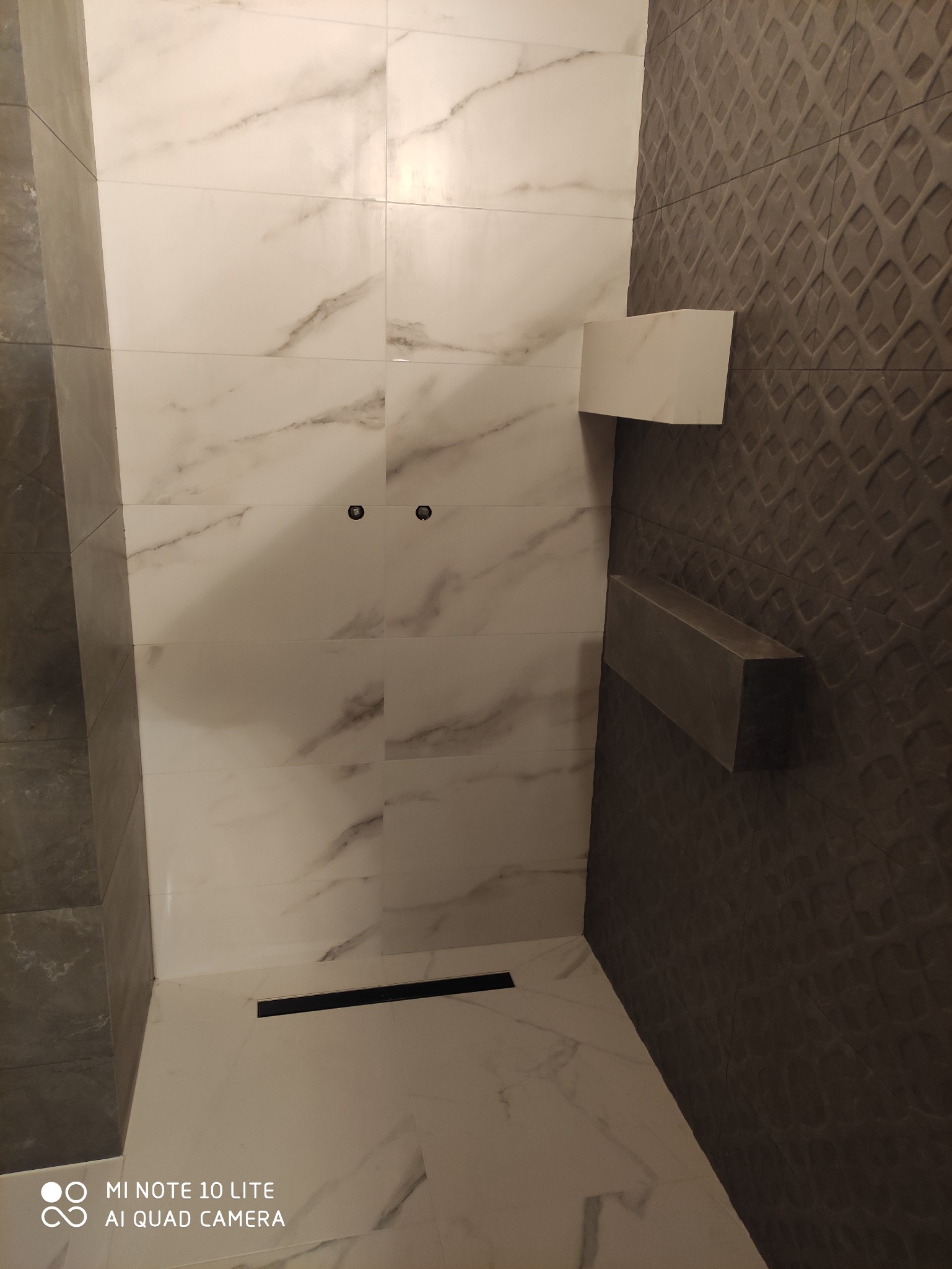 Wykończona kabina prysznicowa z białymi płytkami imitującymi marmur na ścianach i podłodze, czarny odpływ liniowy, dwie półki w kolorze szarego betonu na ścianie z ciemnoszarymi płytkami o fakturze...