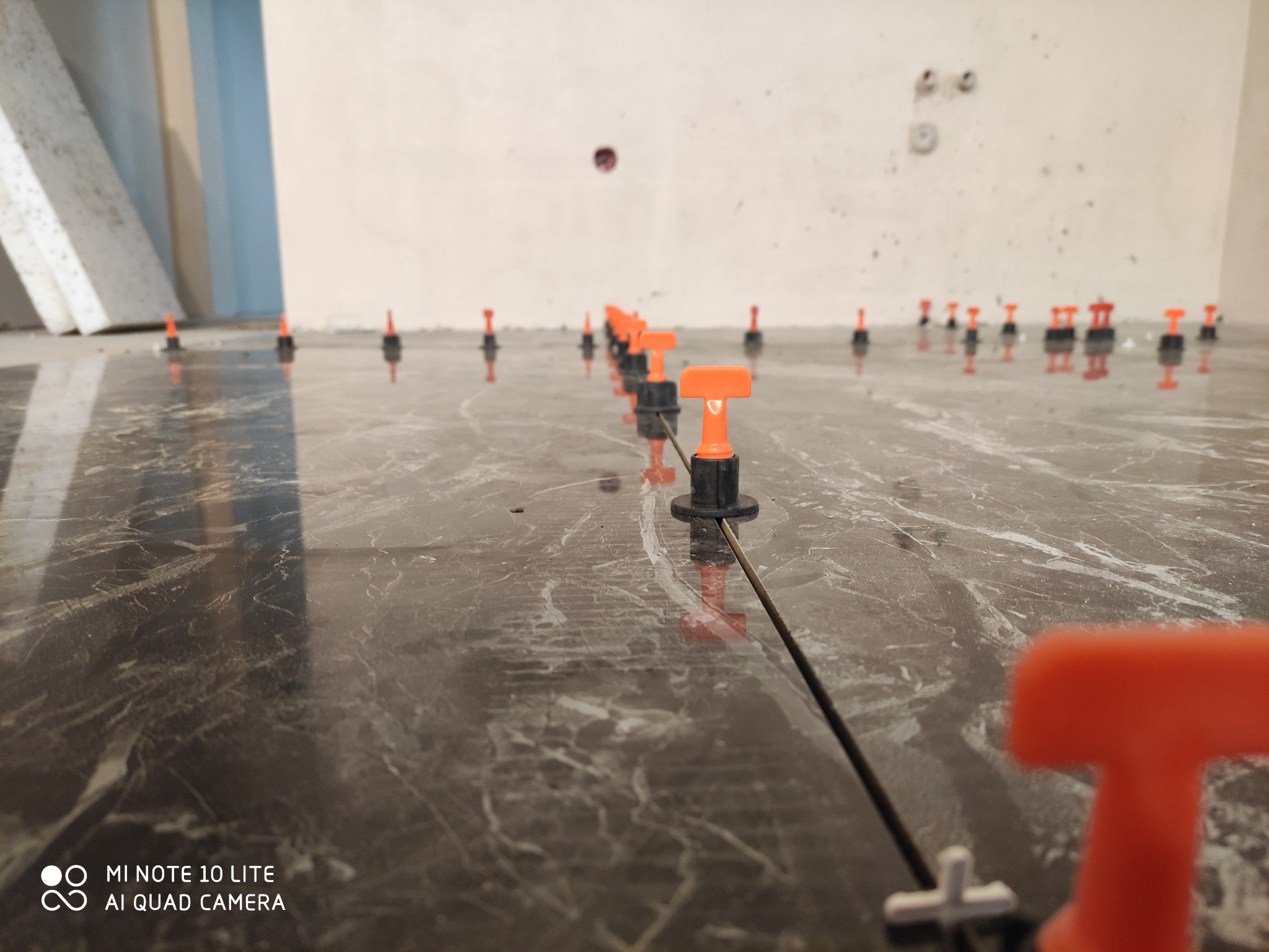 Położona, błyszcząca, marmurkowa płytka podłogowa z pomarańczowymi klinami poziomującymi i krzyżykami dystansowymi w trakcie montażu.