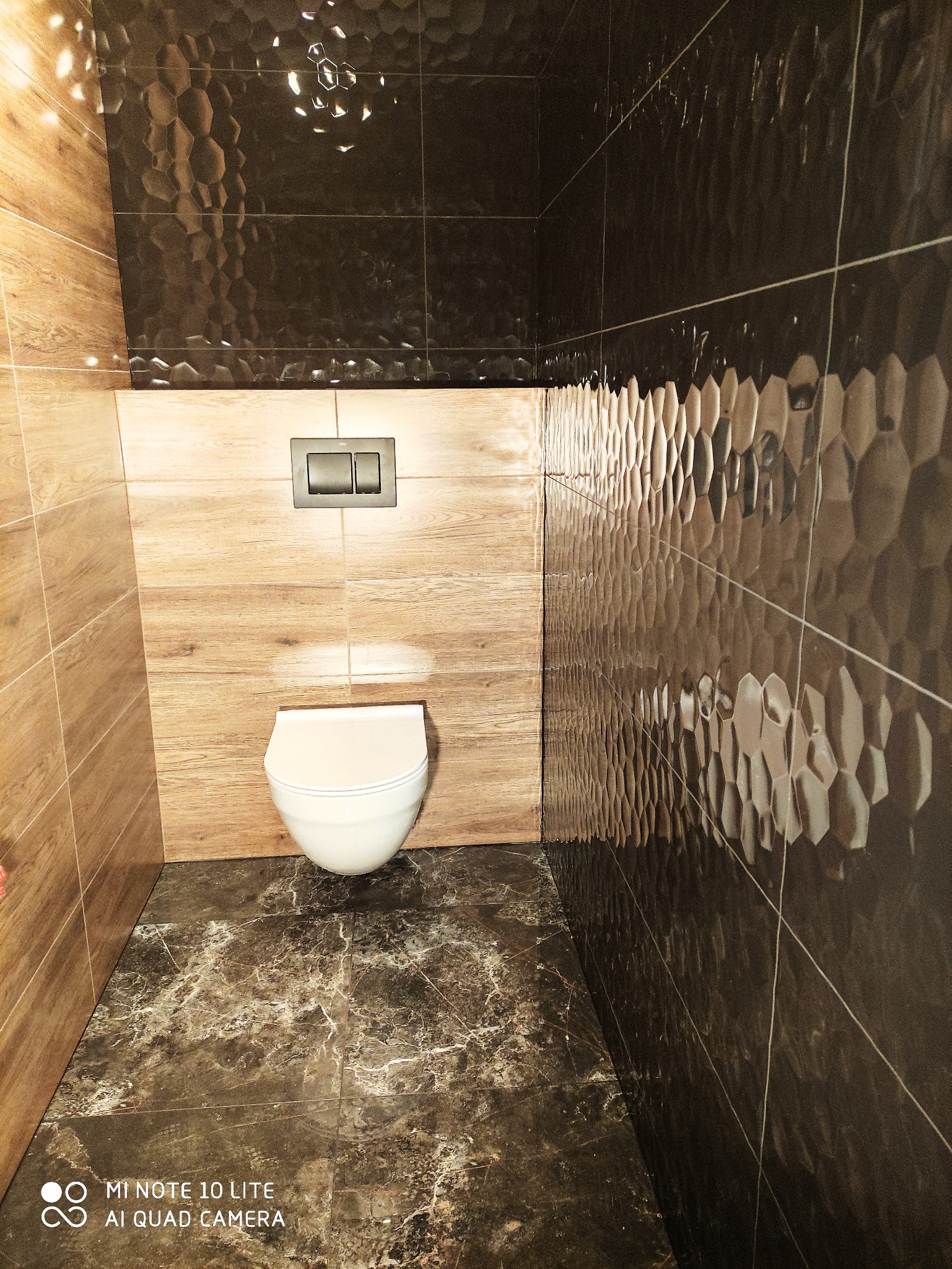 Nowoczesna toaleta podwieszana z szarym przyciskiem spłukującym, drewnopodobne i heksagonalne płytki ścienne, marmurowa podłoga