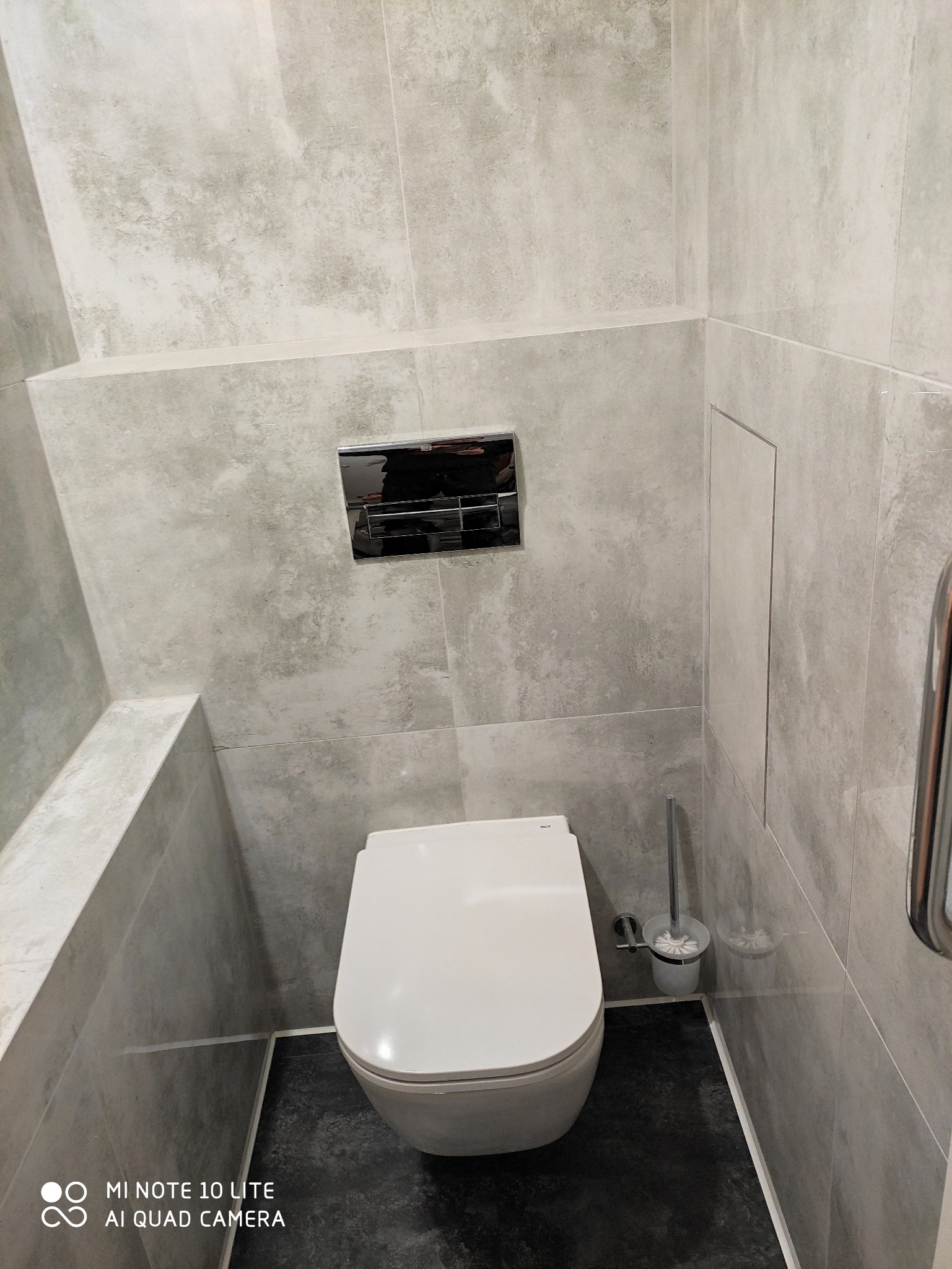 Nowoczesna toaleta wisząca w łazience wykończonej szarymi płytkami imitującymi kamień, z chromowanym przyciskiem spłukującym i szczotką do WC.