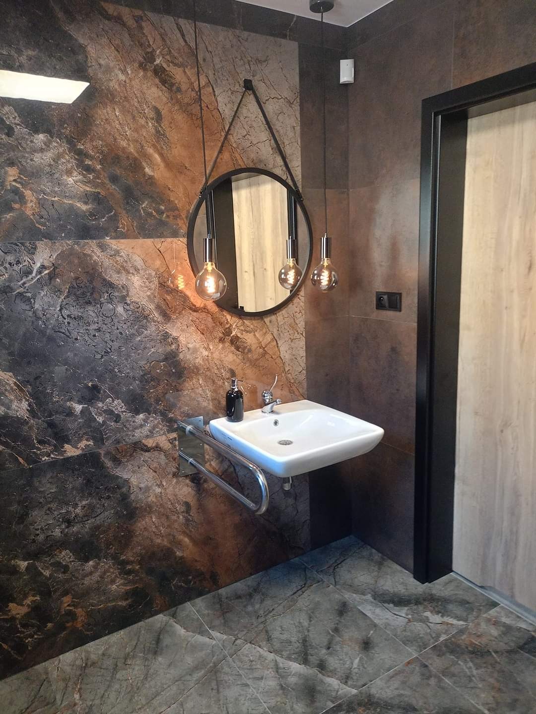 Nowoczesna łazienka z marmurowymi płytkami na ścianach i podłodze, okrągłe lustro z zawieszeniem na pasku, wisząca umywalka z chromowanym wspornikiem i trzema żarówkami na kablach.