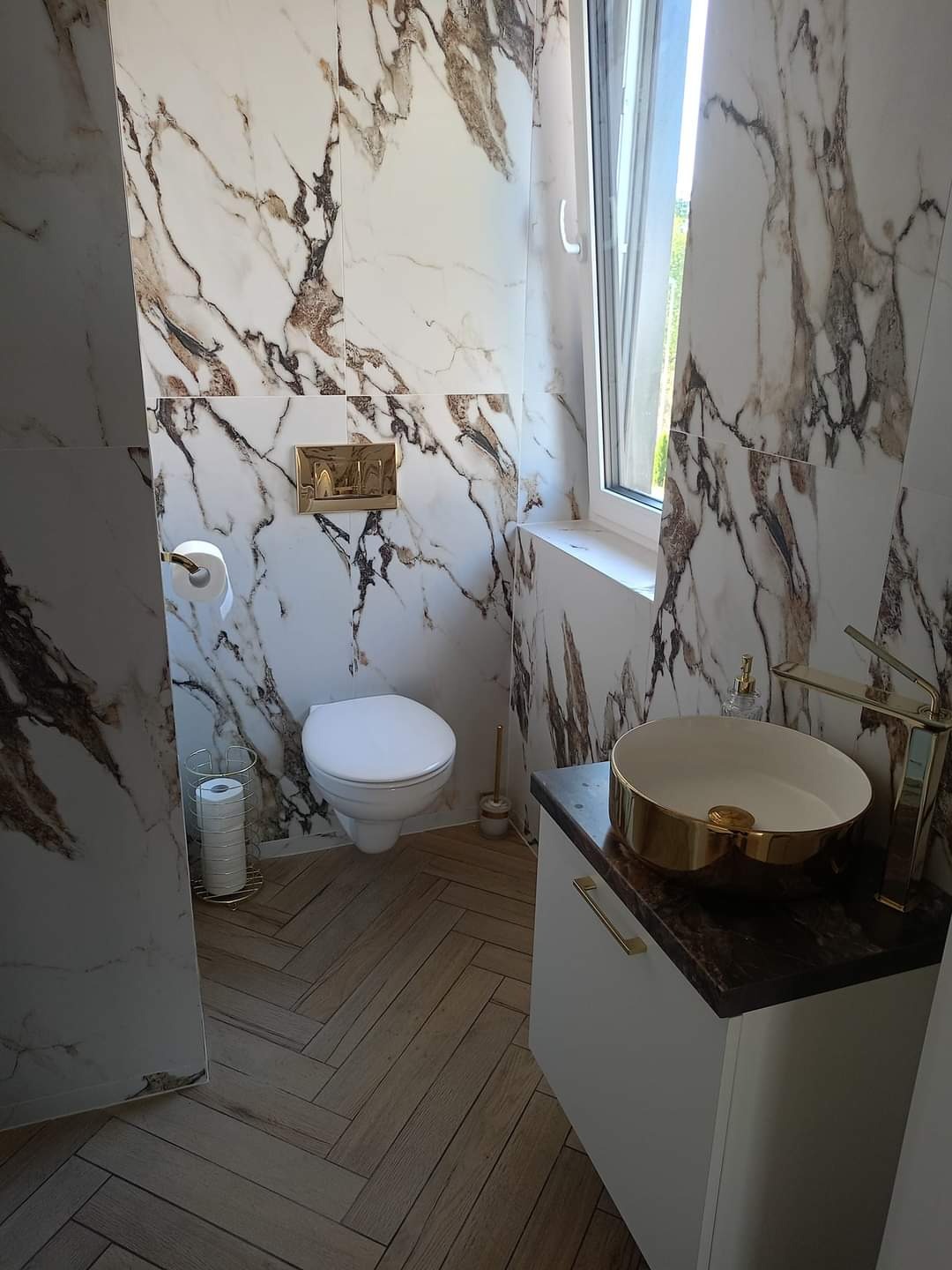 Nowoczesna łazienka z marmurowymi płytkami na ścianach, złotą armaturą i drewnianą podłogą ułożoną w jodełkę.