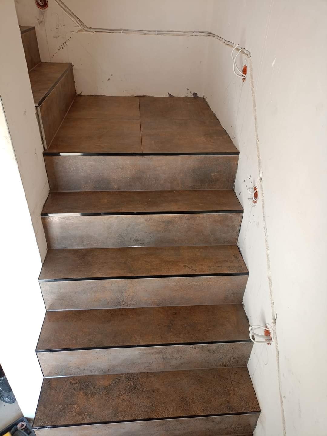 Wykończone płytkami schody wewnętrzne z brązowymi stopnicami i czarnymi paskami na krawędziach, widoczne kable i puszki elektryczne na ścianach w stanie surowym.