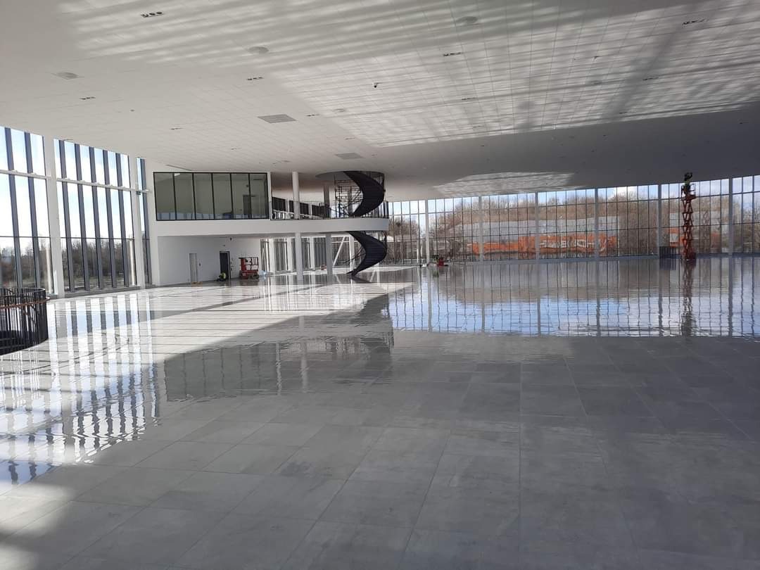 Przestronne wnętrze biurowe z szarą, wypolerowaną podłogą, spiralnymi schodami i dużymi oknami z widokiem na zewnątrz, widoczny podnośnik koszowy.