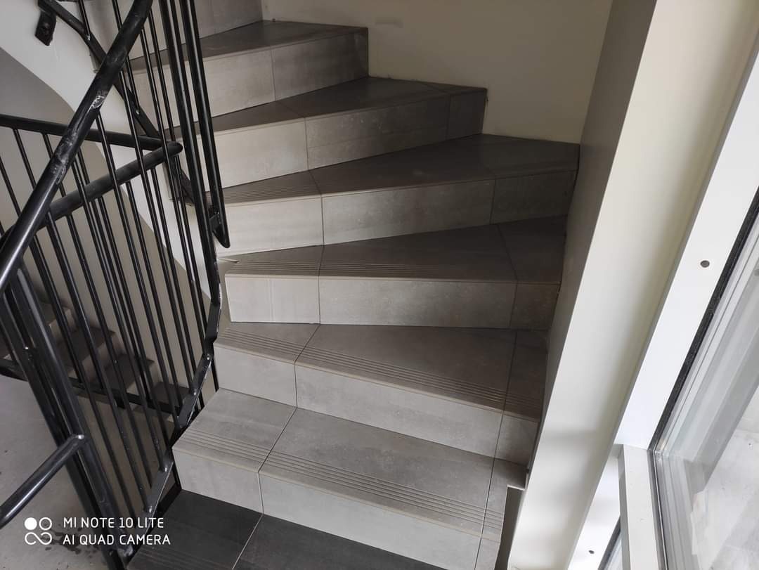 Schody spiralne wyłożone szarymi płytkami ceramicznymi z metalową balustradą, widok z góry, neutralne oświetlenie.