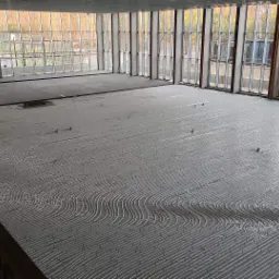 Szare rurki ogrzewania podłogowego ułożone w faliste wzory na dużej powierzchni podłogi w trakcie instalacji, widoczne metalowe ramy okienne i betonowa wylewka w tle.