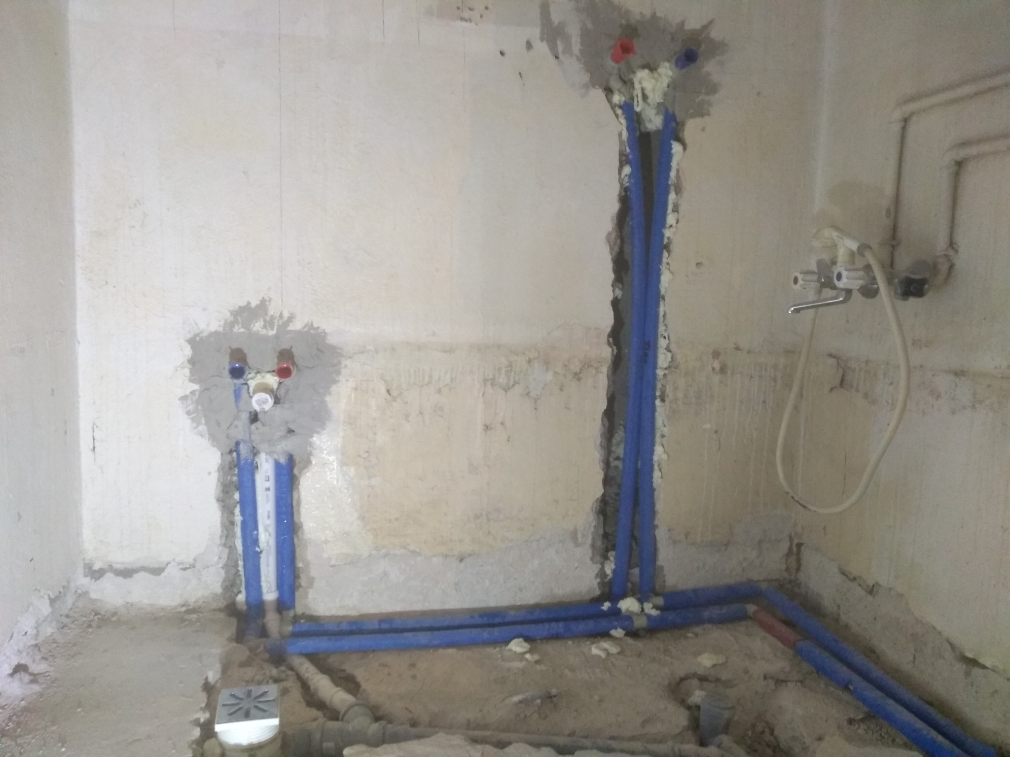 Surowe wnętrze łazienki w trakcie prac instalacyjnych: widoczne nieotynkowane ściany, nowe niebieskie rury wodne zabezpieczone pianką montażową, zamontowana bateria umywalkowa i odpływ podłogowy...
