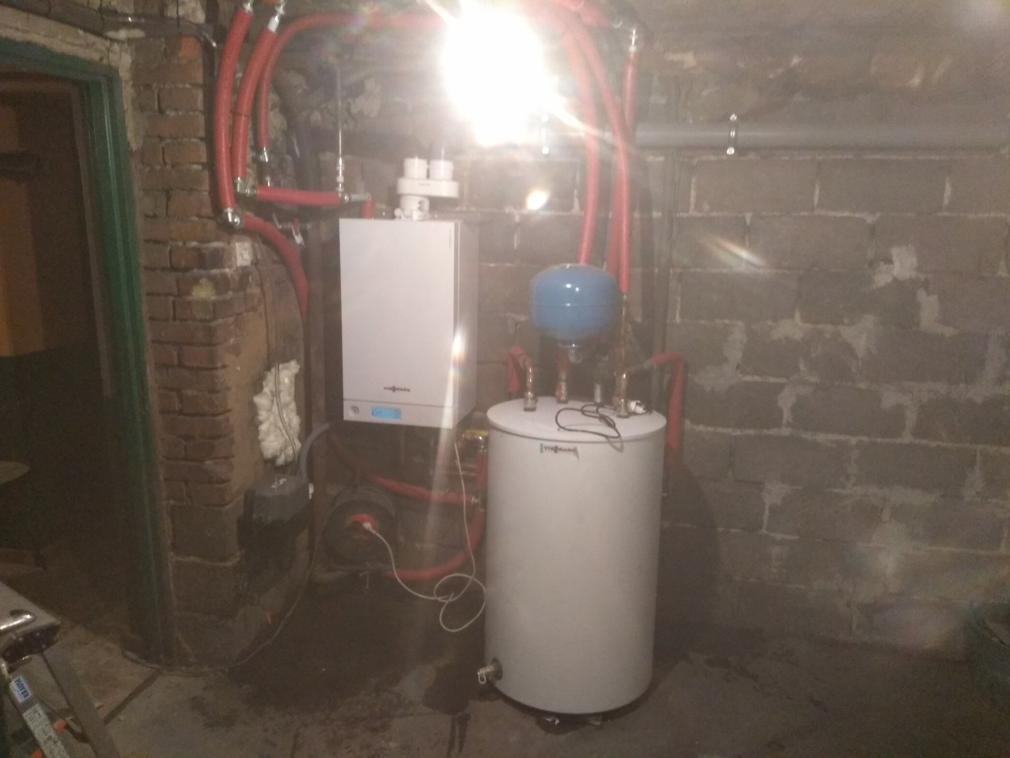 Nowoczesny piec grzewczy Viessmann z zasobnikiem wody, połączony czerwonymi rurami w pomieszczeniu z surowymi, ceglanymi ścianami i betonową podłogą. Widoczne elementy instalacji i fragment drabiny.