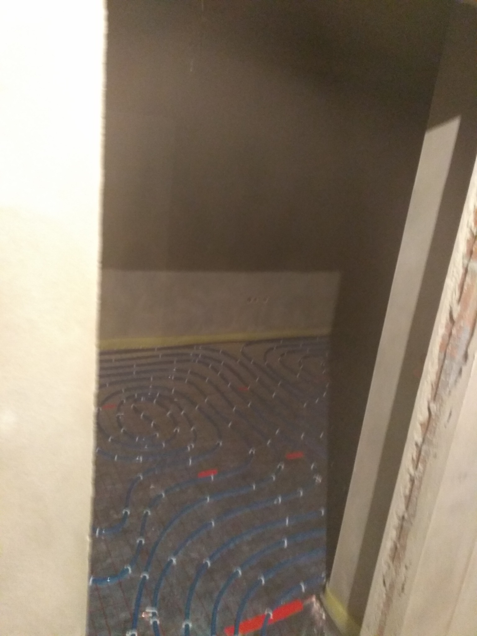 Niedokończona instalacja ogrzewania podłogowego z widocznymi niebieskimi rurkami i czerwonymi elementami mocującymi, częściowo zalana szlichtą, widok z góry z perspektywy wnęki w ścianie.