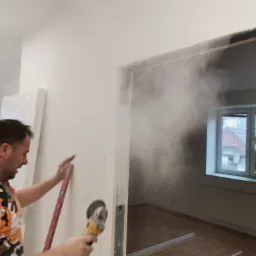 Pracownik z szlifierką kątową podczas wycinania otworu w ścianie, unoszący się pył, poziomica oparta o ścianę, widoczne elementy ościeżnicy i okno w tle.