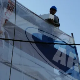 Pracownik w niebieskim kasku, stojący na rusztowaniu osłoniętym plandeką z nadrukiem, trzyma białą płytę styropianową na tle błękitnego nieba.