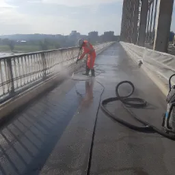 Pracownik w pomarańczowym kombinezonie i kasku czyści nawierzchnię mostu za pomocą myjki ciśnieniowej; w tle widoczna panorama miasta.