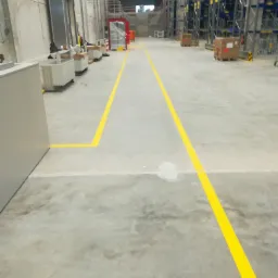 Magazynowa posadzka betonowa z wyznaczonymi żółtymi liniami komunikacyjnymi, widoczne regały w tle.