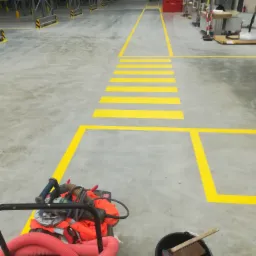 Oznakowanie żółtą farbą na posadzce w hali magazynowej, widoczne linie, przejście dla pieszych oraz wózek z narzędziami i wiadro z akcesoriami malarskimi.