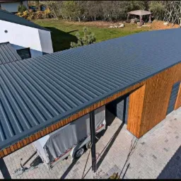 Nowoczesny, dwuspadowy dach z blachy trapezowej w kolorze grafitowym, połączony z drewnianą konstrukcją w stylu skandynawskim, widok z góry, na podjeździe zaparkowana przyczepa z plandeką.