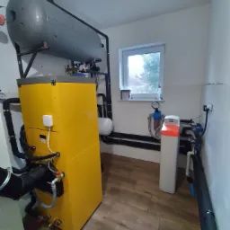 Żółty piec na paliwo stałe z szarym zbiornikiem akumulacyjnym w kotłowni z oknem i zmiękczaczem wody.