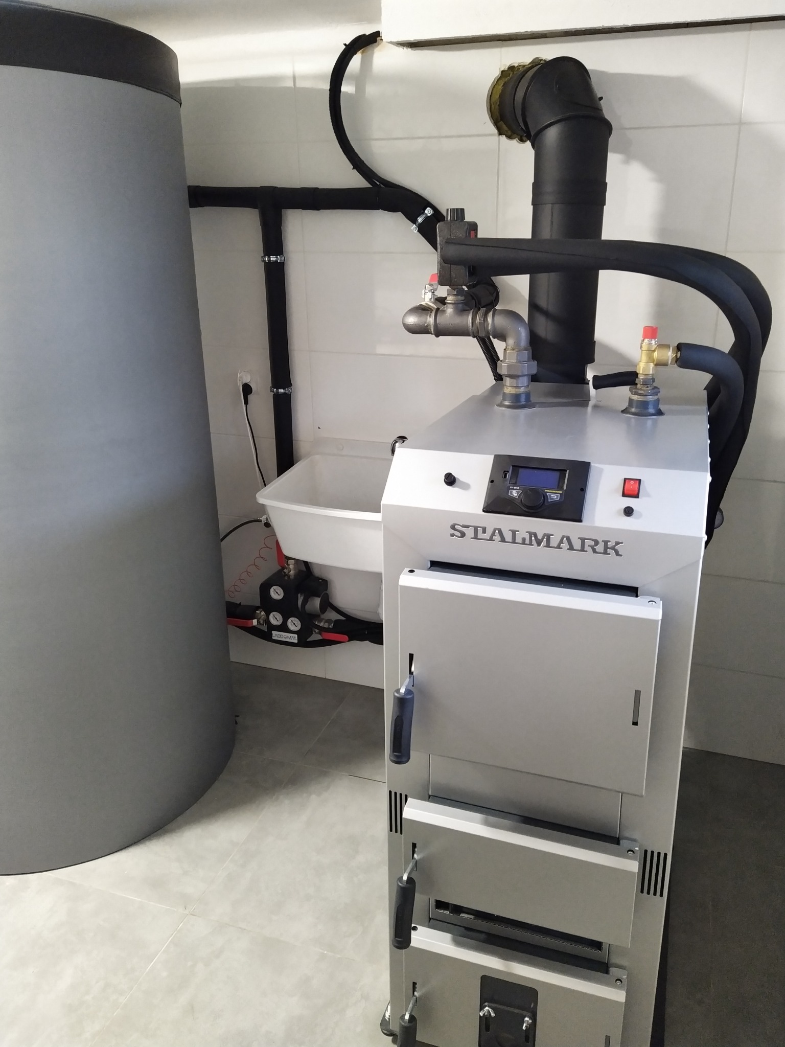 Nowoczesny piec marki STALMARK z podajnikiem, zainstalowany w kotłowni z białymi płytkami, widoczny zbiornik akumulacyjny i elementy instalacji.