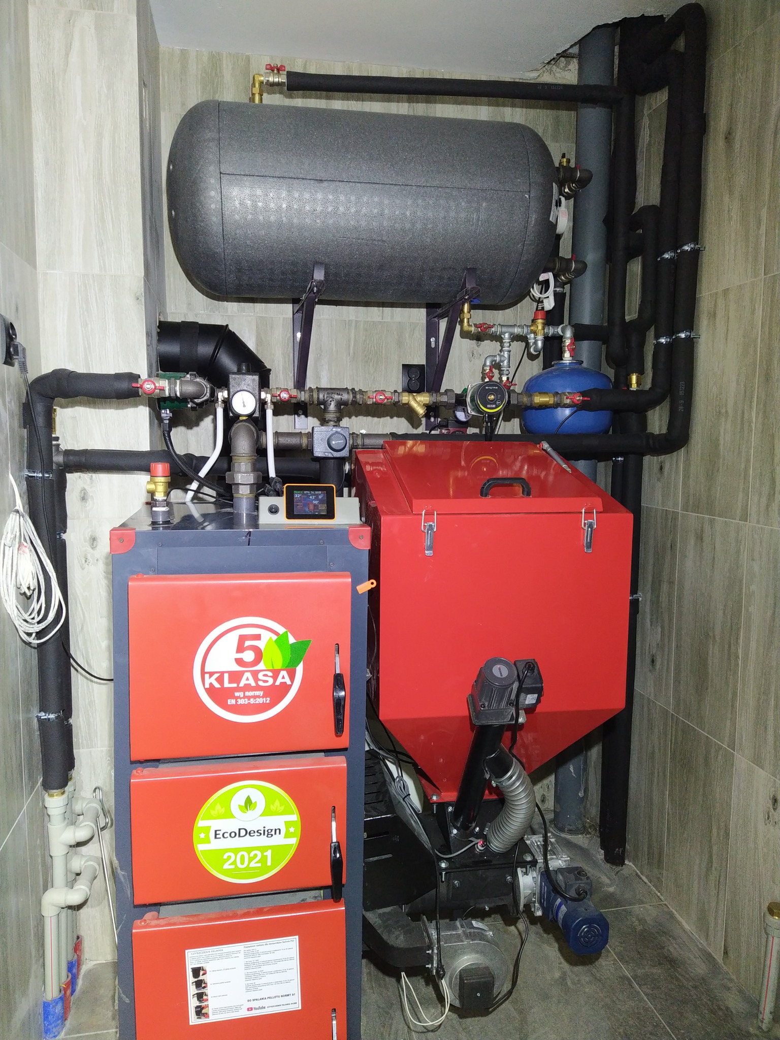 Nowoczesny piec na paliwo stałe z zasobnikiem i instalacją, widoczne oznaczenia klasy 5 i EcoDesign 2021, zainstalowany w kotłowni z wykończeniem imitującym drewno.