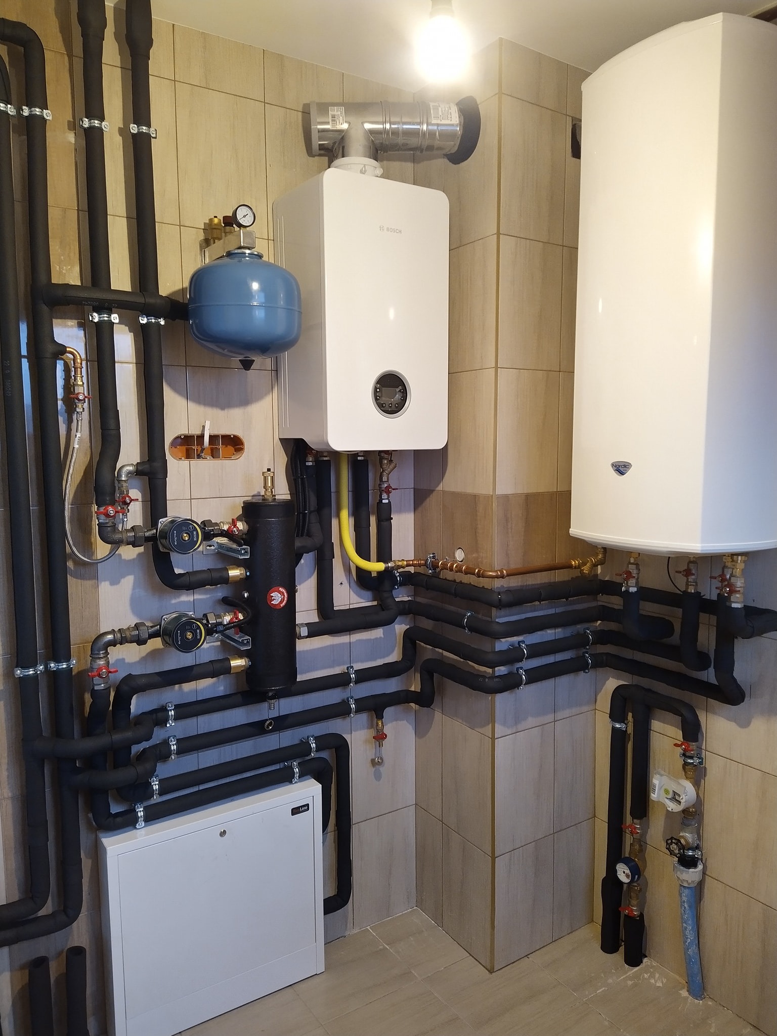Zrealizowana instalacja centralnego ogrzewania z piecem gazowym Bosch, zbiornikiem buforowym i zasobnikiem wody, rury w izolacji, płytki ceramiczne na ścianach.