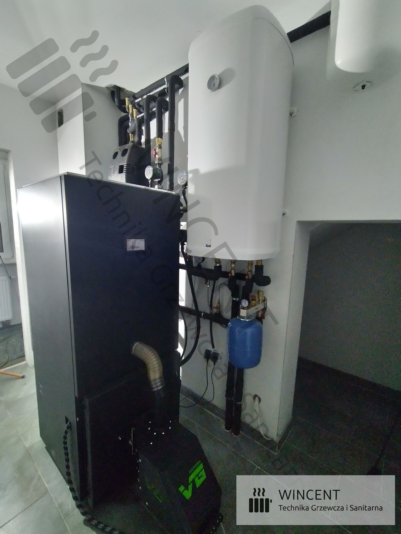 Czarny piec na paliwo stałe z podajnikiem, obok biały zasobnik ciepłej wody, instalacja grzewcza z czarnych rur, niebieski zbiornik przeponowy, szare płytki podłogowe.