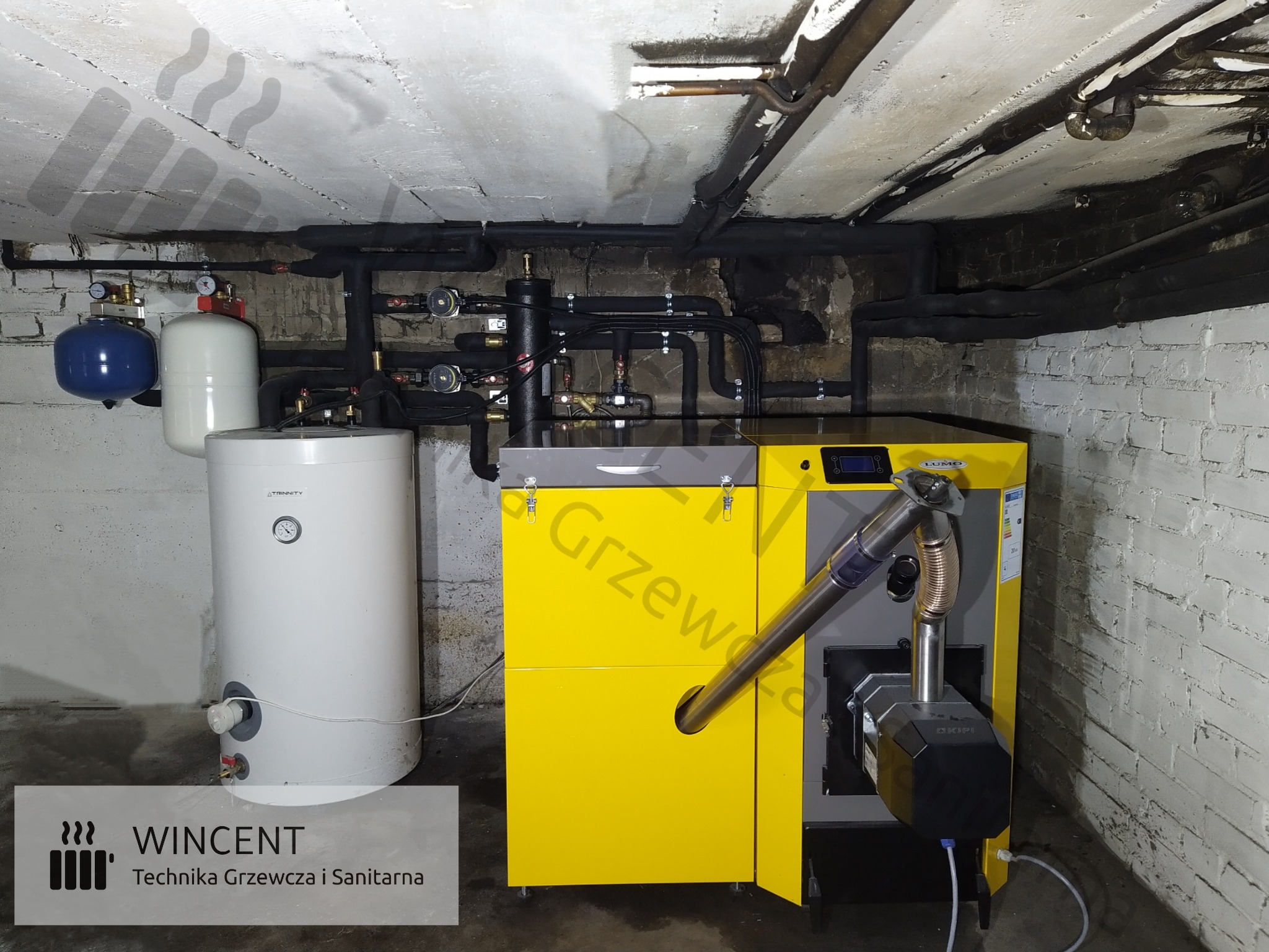 Żółto-szary piec z podajnikiem w kotłowni z instalacją, zbiornik wyrównawczy obok, rury izolowane na suficie, ściany z cegły pomalowane na biało.