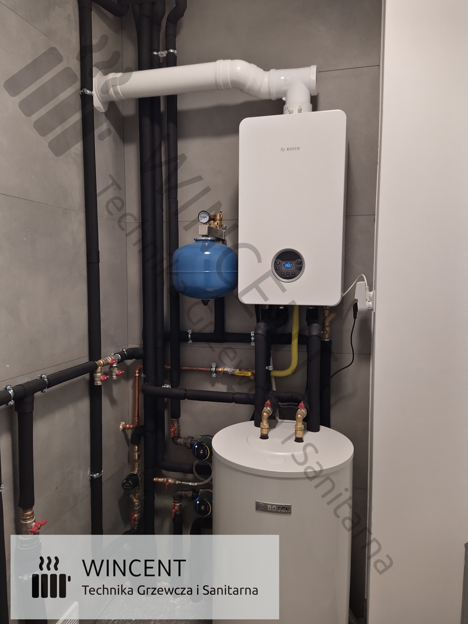 Biały, kondensacyjny kocioł gazowy Bosch z podgrzewaczem wody, niebieskim naczyniem wzbiorczym i czarnymi rurami w szarej kotłowni.