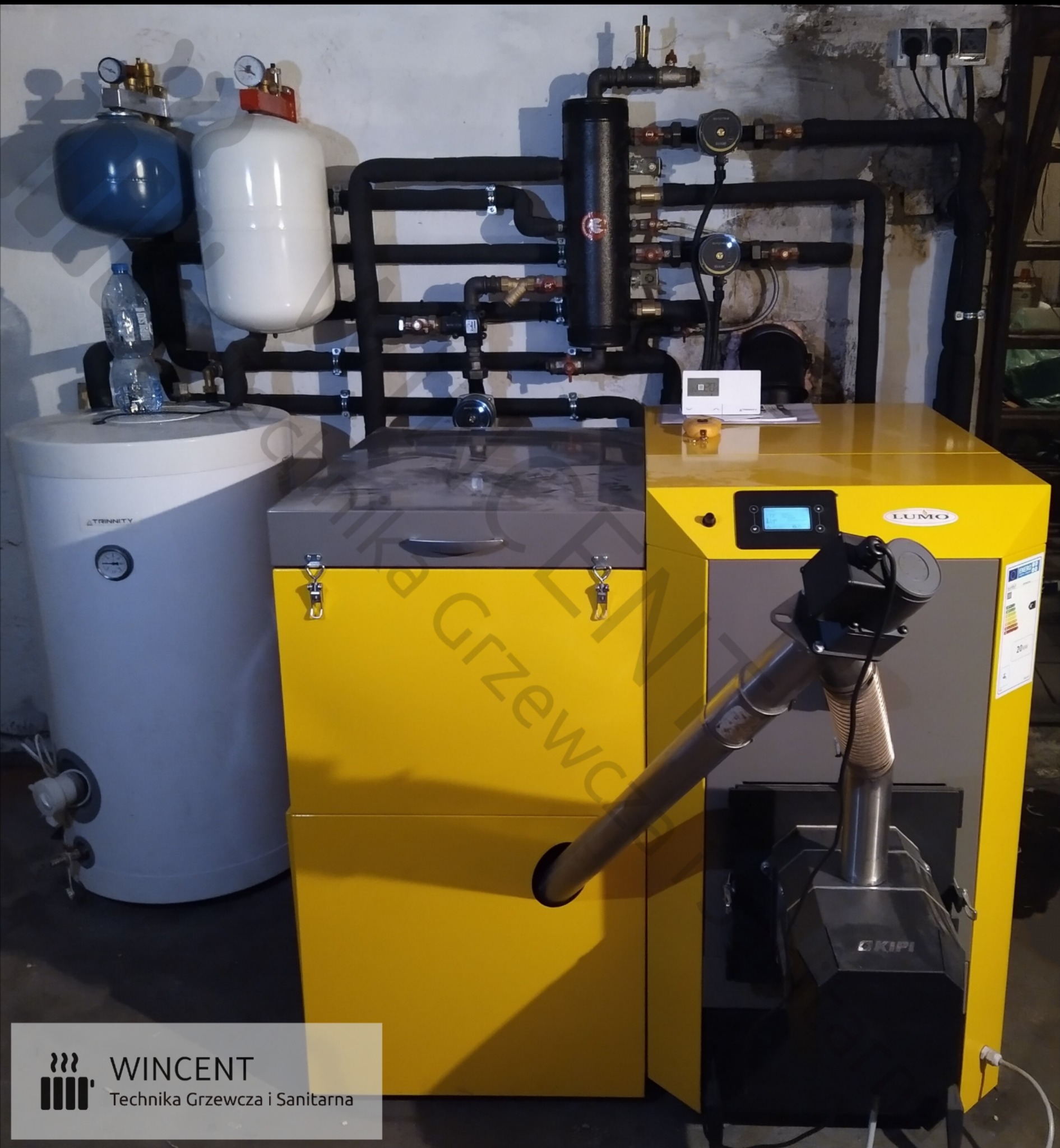 Żółto-szary piec na paliwo stałe LUMO z podajnikiem Kipi, widoczne rury instalacji grzewczej i zbiornik buforowy Trinity, butelka wody na zbiorniku.