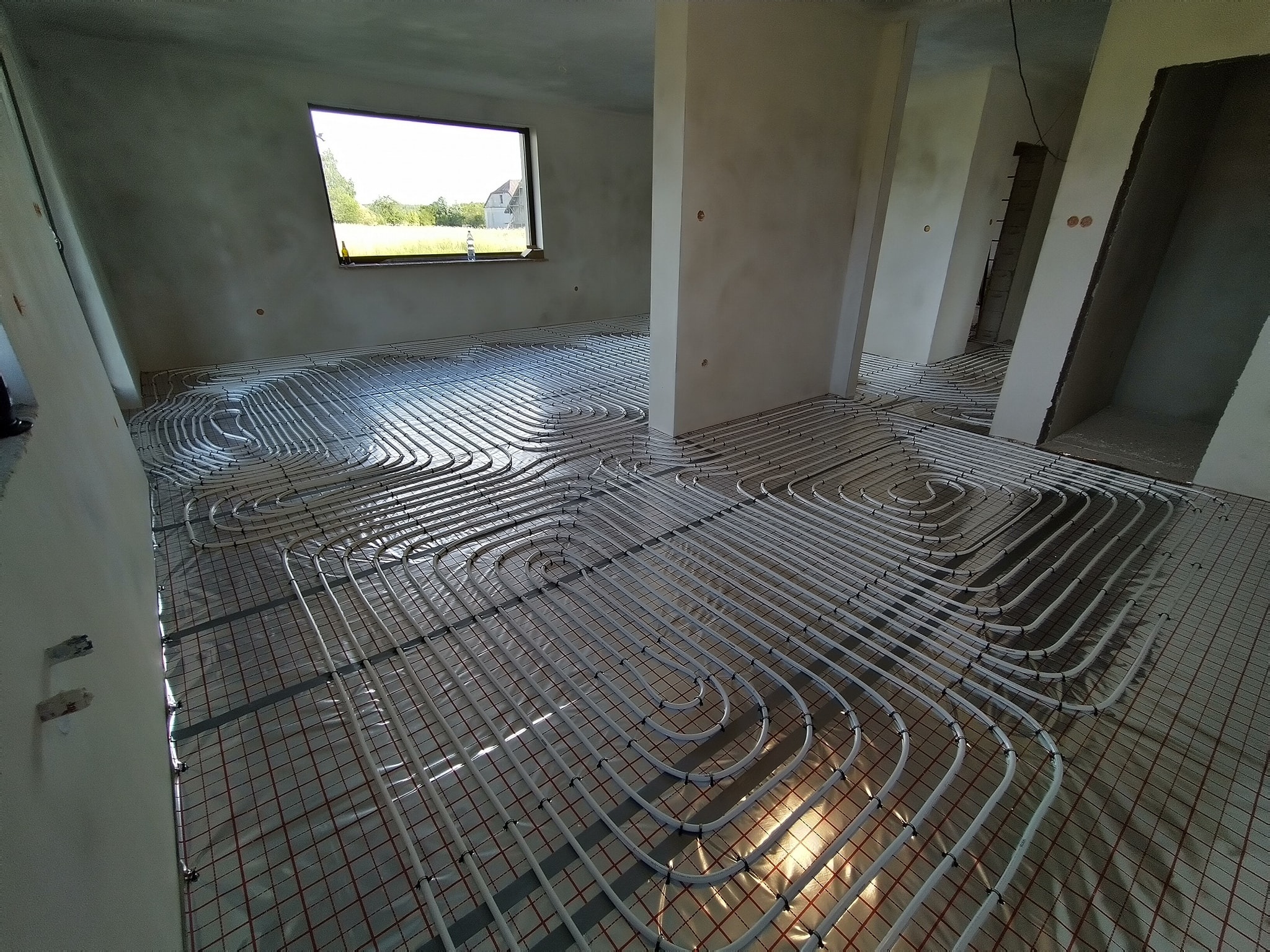 Białe rury ogrzewania podłogowego ułożone spiralnie na folii z siatką w surowym wnętrzu domu z oknem.