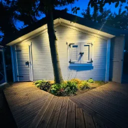 Oświetlona altana z białymi ścianami i drewnianym tarasem otaczającym drzewo, widoczna wieczorem z podświetleniem punktowym.